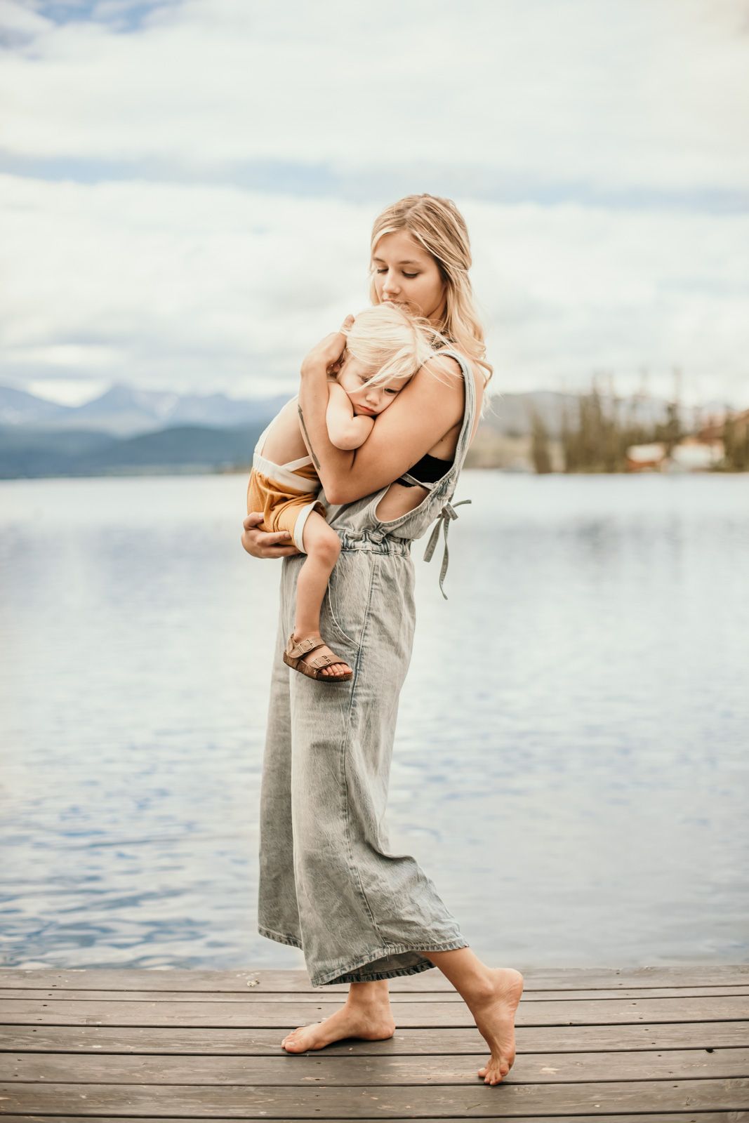 A parent and child share a tender embrace while standing barefoot on a wooden dock by a serene mountain lake.
