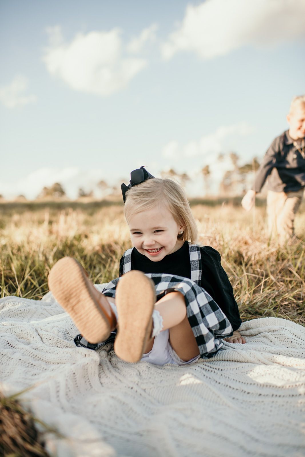 A child wearing a black and white plaid dress lies on a white blanket in a sunlit field of tall grass.