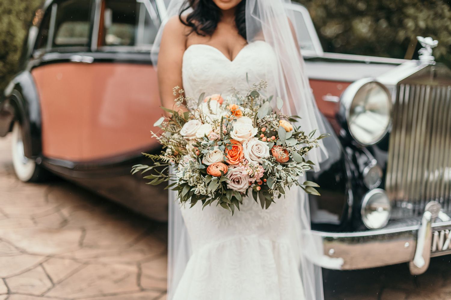 A beautiful wedding bouquet with peach and white flowers sits alongside vintage cars at an elegant outdoor venue.