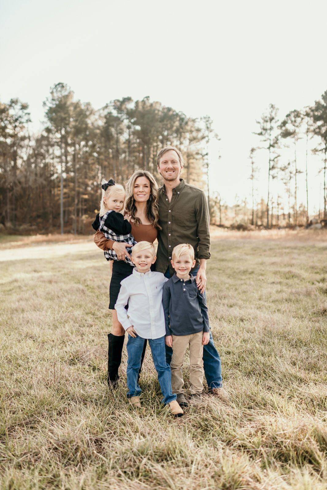 A family poses together in a grassy field with tall pine trees in the background during golden hour.