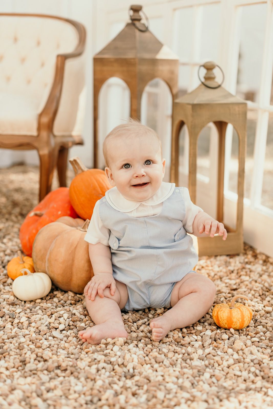 A baby in grey clothing sits among autumn pumpkins and decorative lanterns.