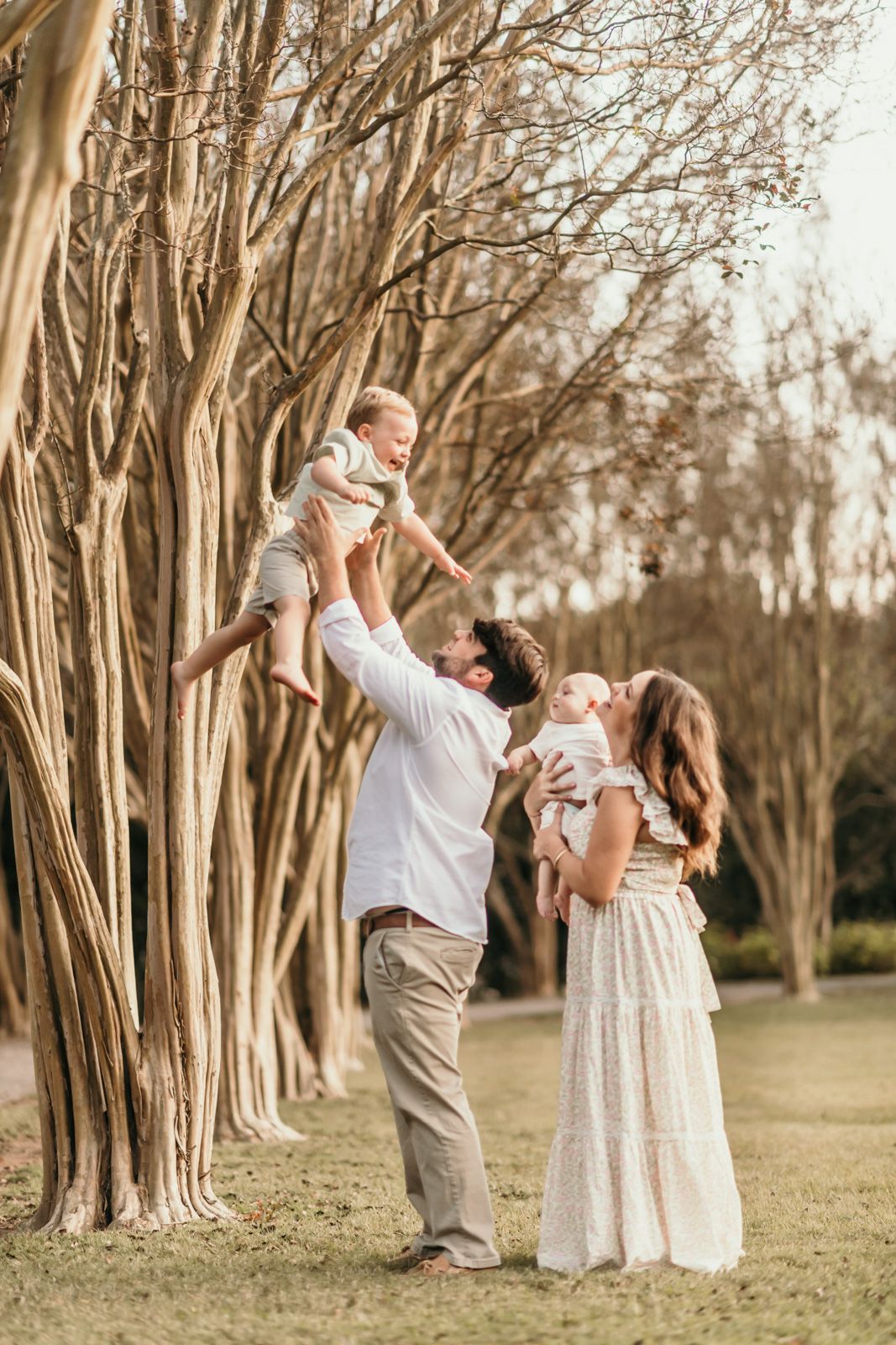 A couple in white and beige outfits plays with their baby in a scenic grove of trees at sunset.