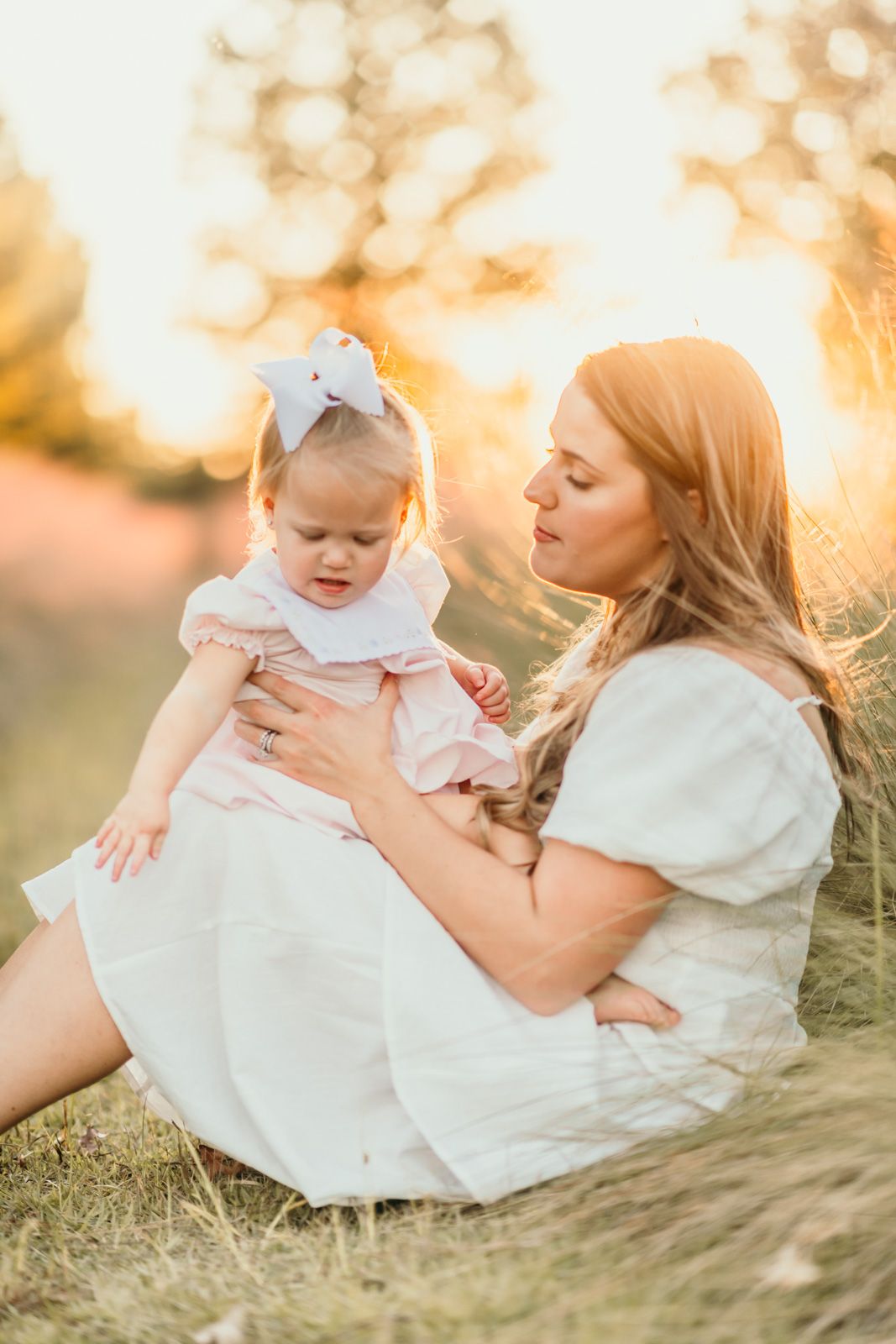 A tender moment captured at sunset as a parent and baby in white dresses sit together in a grassy meadow.