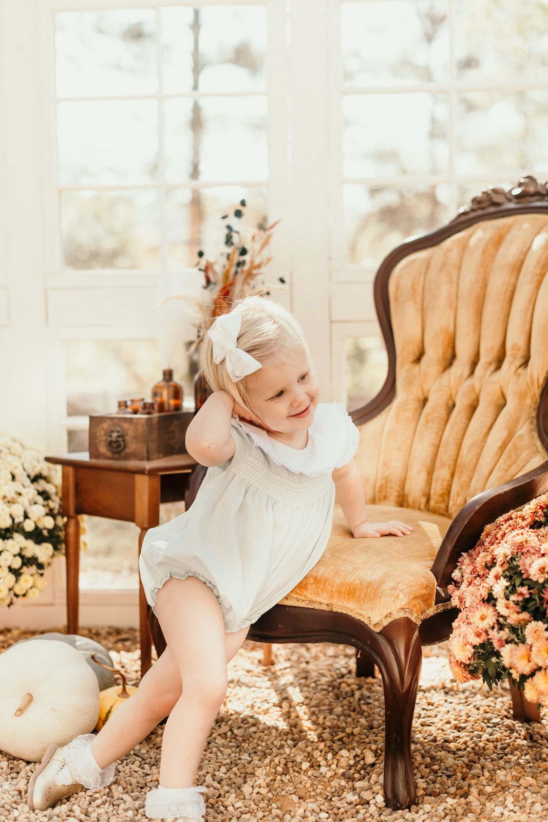 A small child plays on a vintage velvet chair next to autumn flowers and decor.