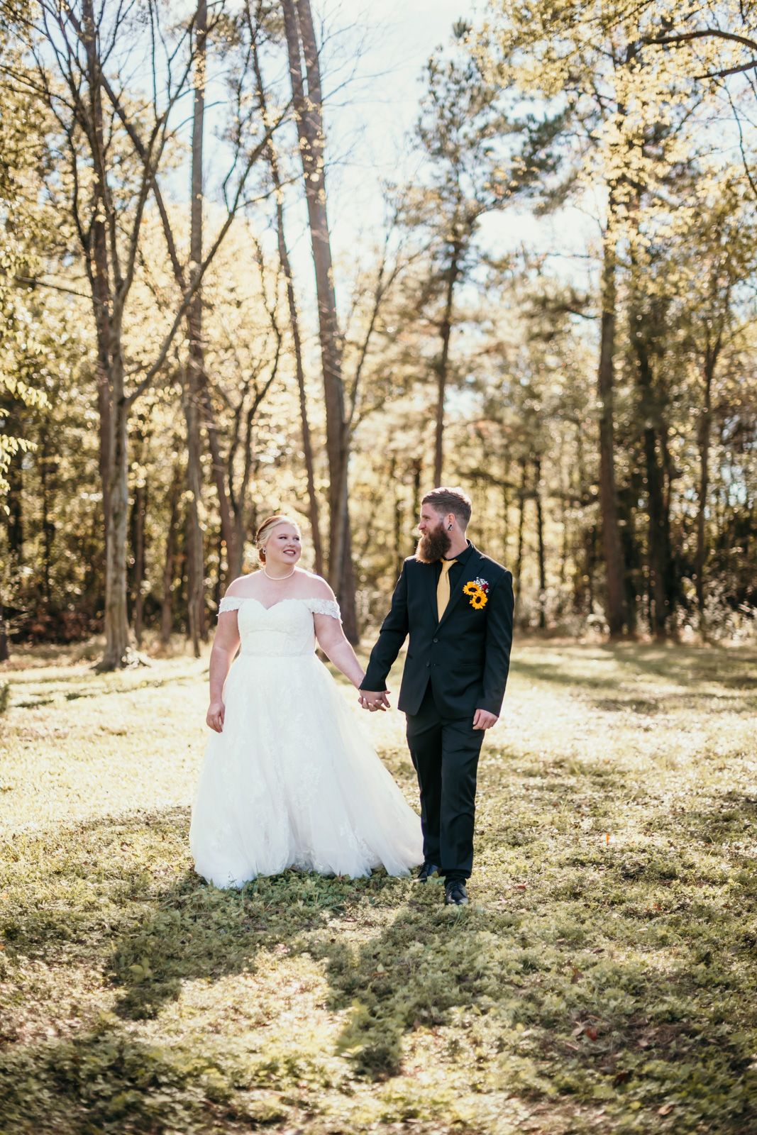 A couple in wedding attire walks hand in hand through a sunlit autumn forest with golden light filtering through trees.