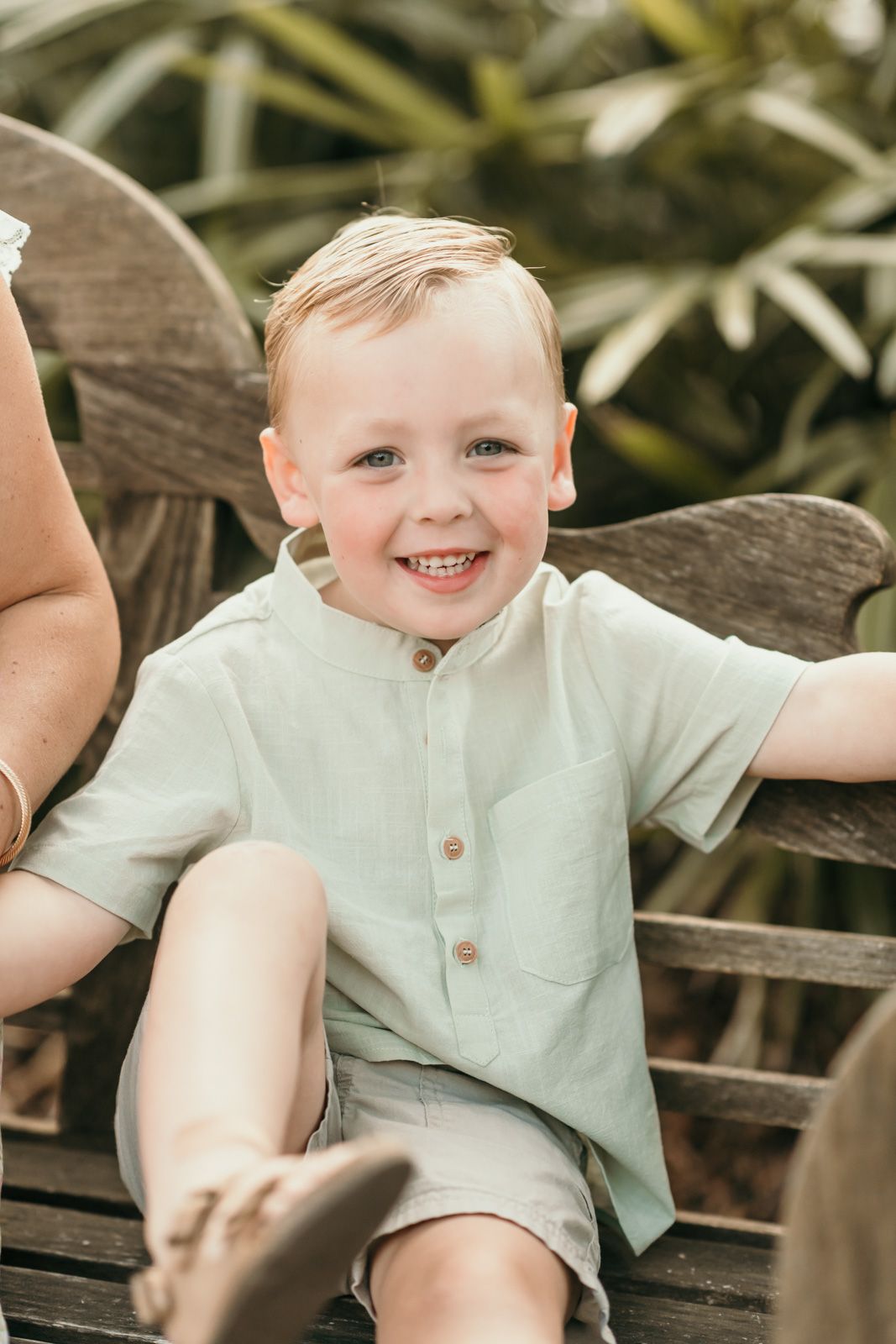 Child in mint green shirt smiles while lounging on wooden bench.
