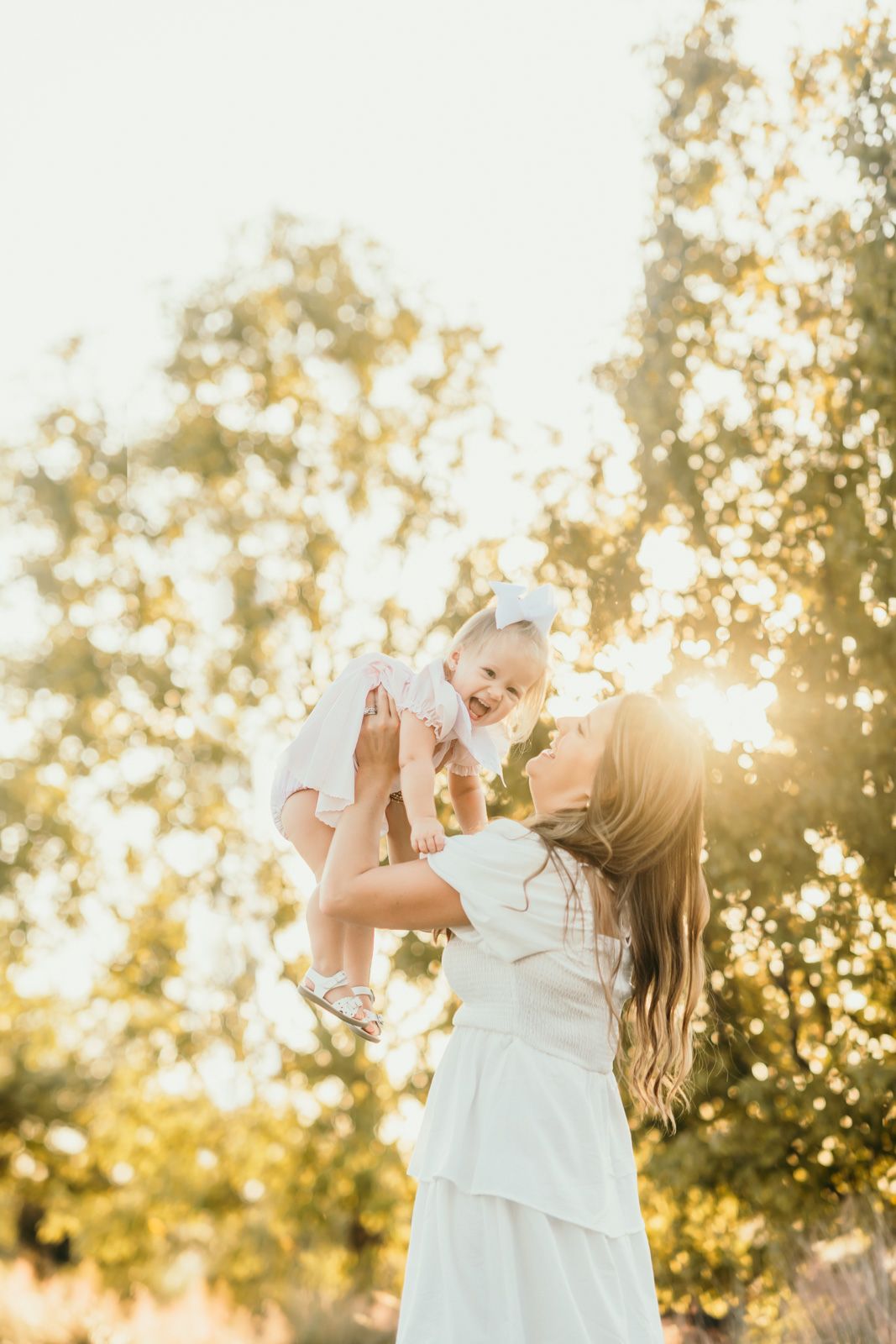 Mother and child playing together in a sunlit garden wearing white dresses during golden hour.