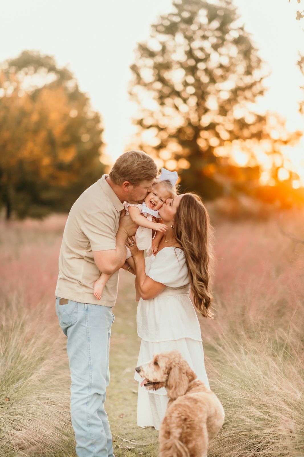 A couple shares a romantic kiss in a golden field at sunset while their fluffy dog watches nearby.