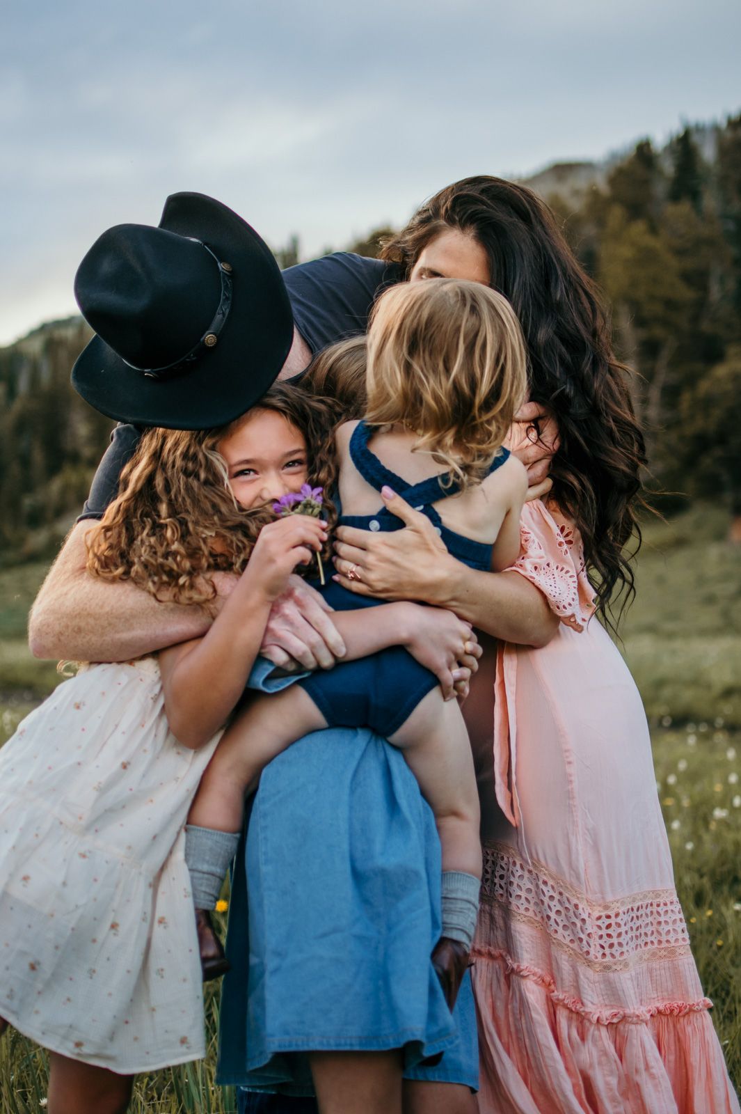 Three children in summer outfits share a sweet group hug together outdoors in a field on a sunny day.