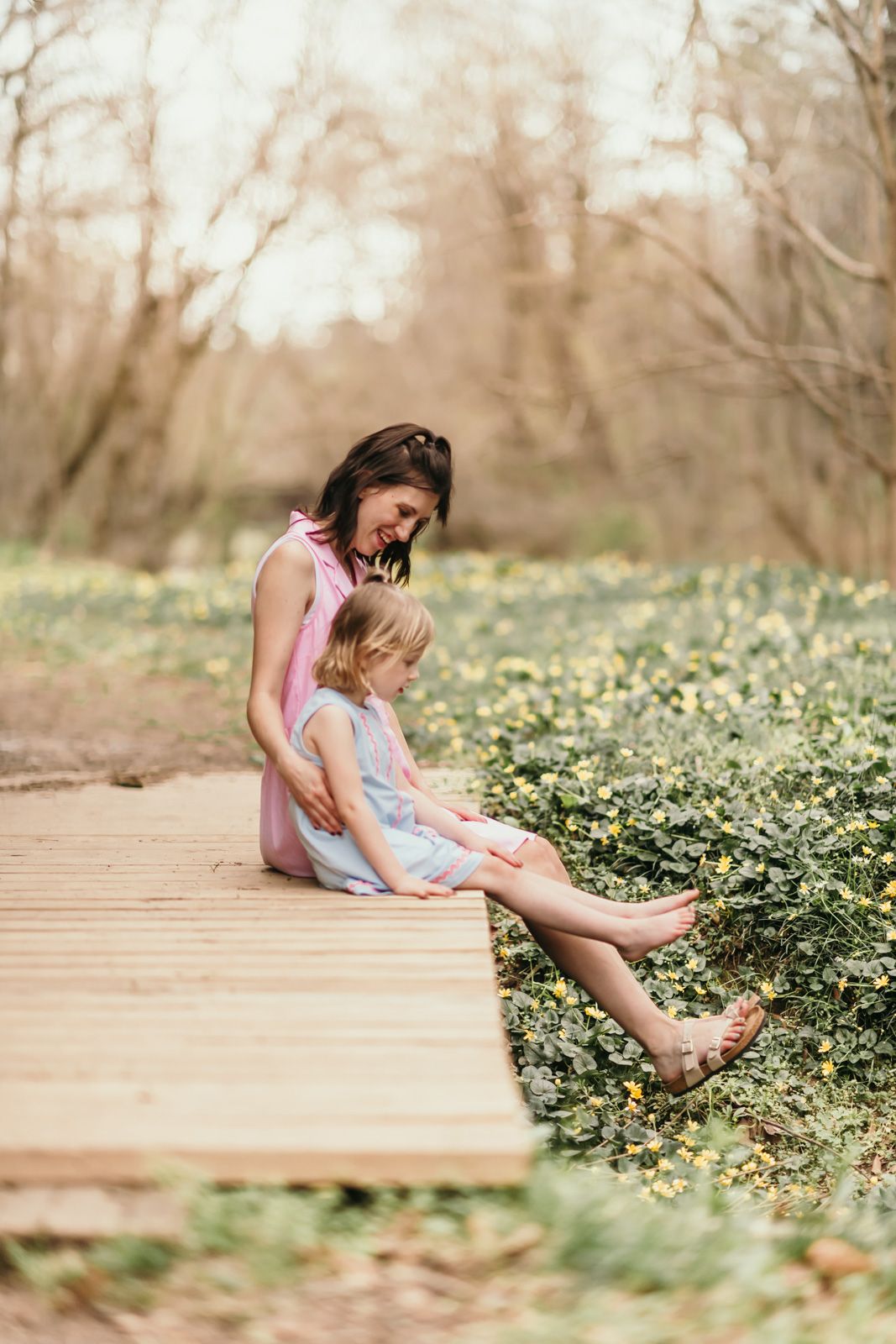 A parent and child sit together on a wooden boardwalk surrounded by blooming white wildflowers on a spring day.