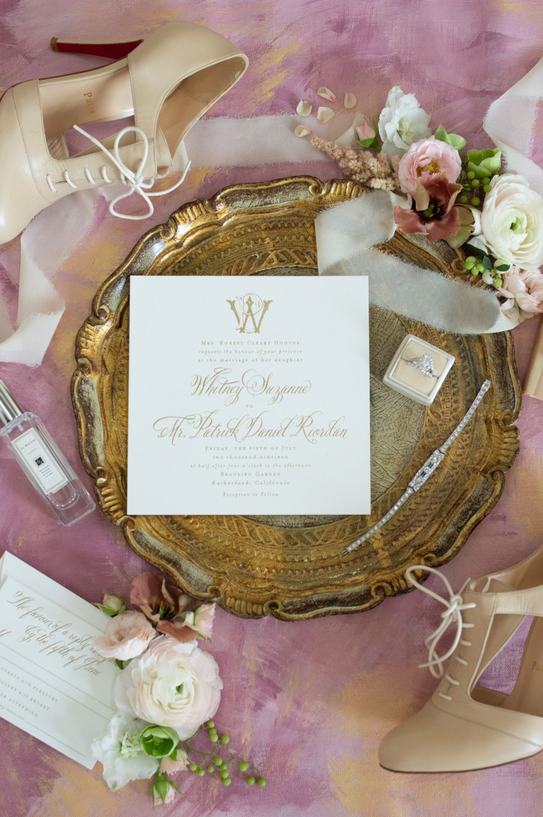 Elegant wedding invitation suite with gold calligraphy on white cards displayed on vintage gold trays with pink flowers.