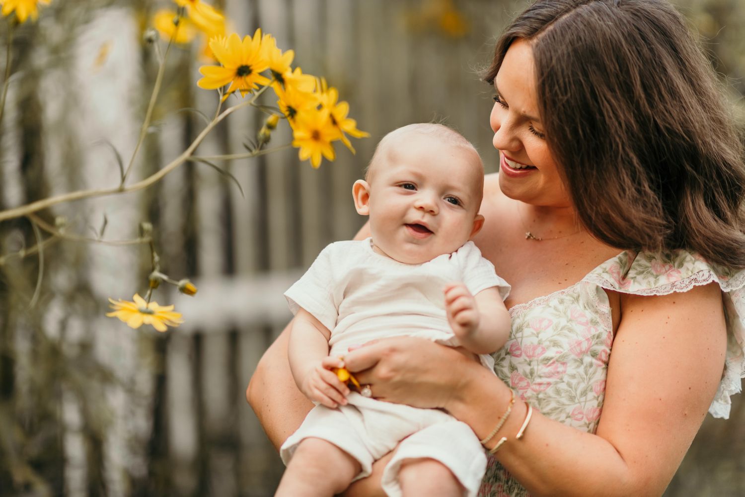 A parent holds a baby outdoors while yellow flowers bloom in the trees above them during a sunny spring day.