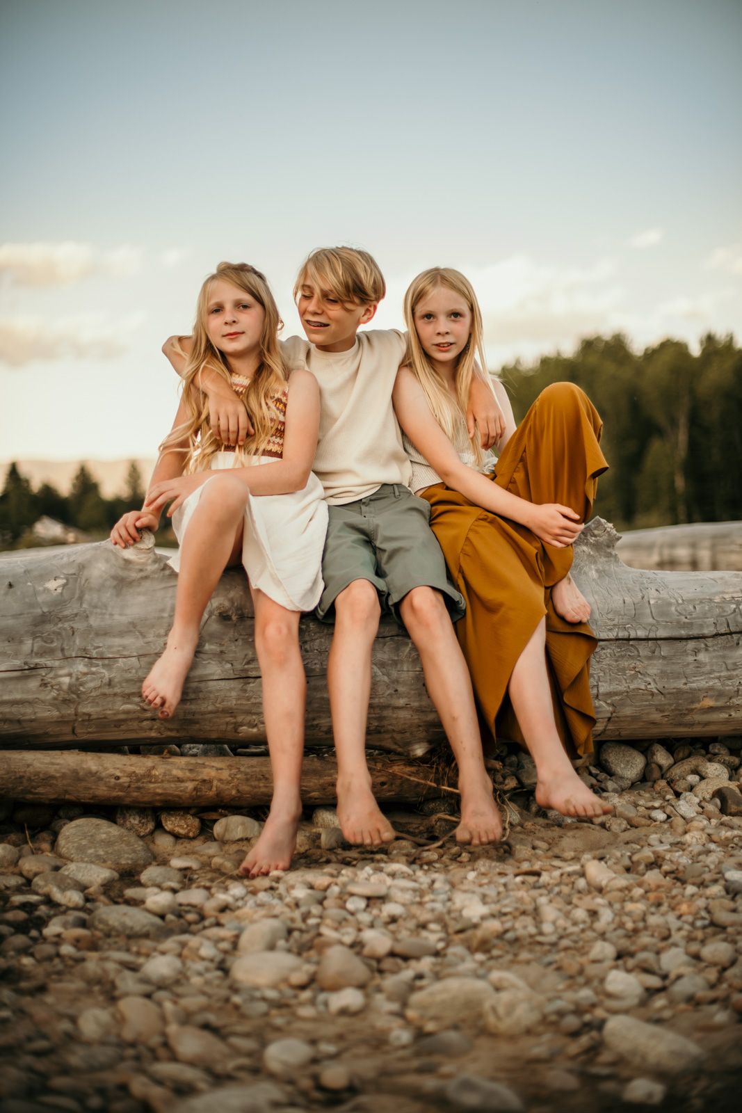 Three children sit barefoot on a large log at sunset with a rocky beach and forest in the background.