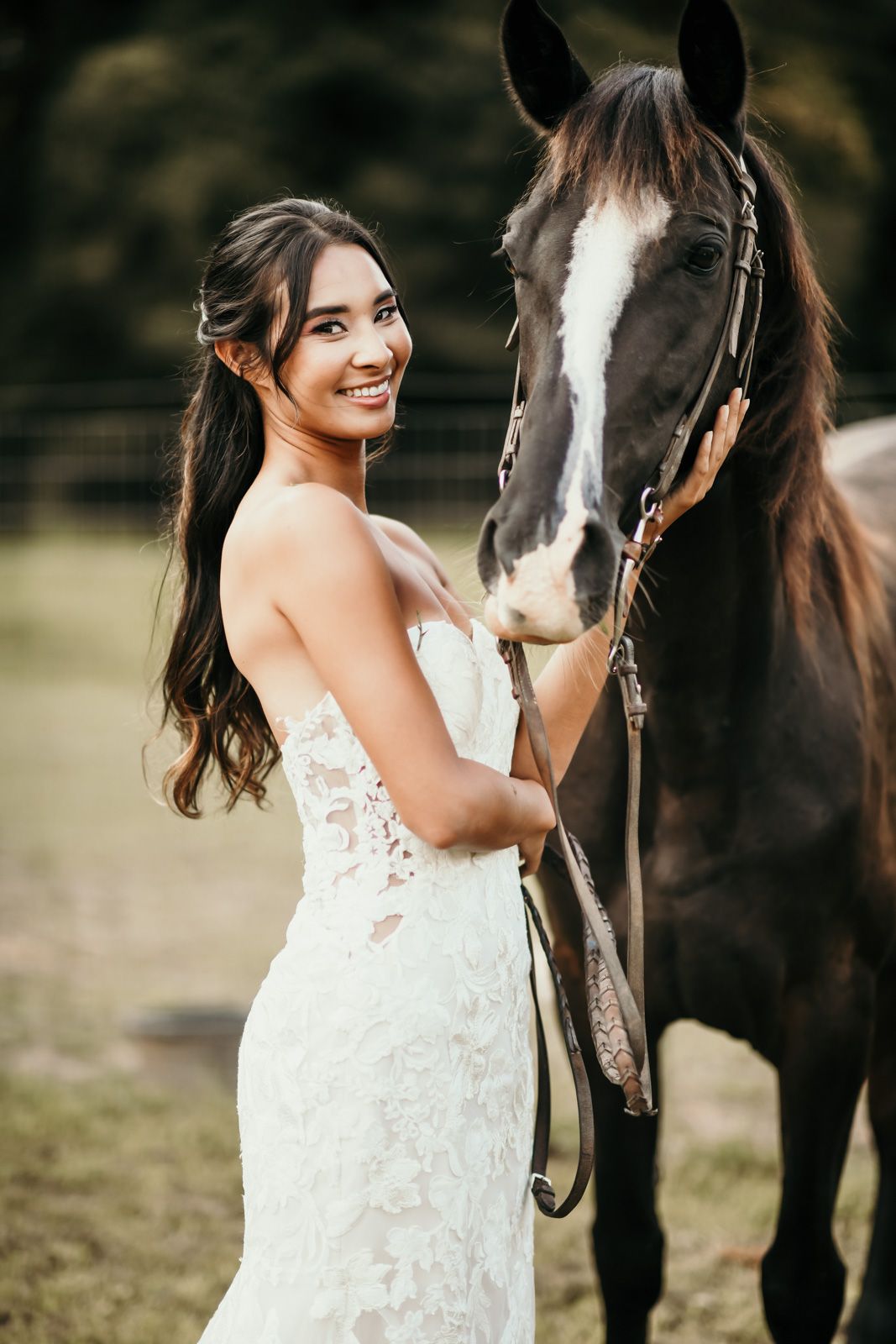 A beautiful bride in a white lace wedding dress poses with a dark brown horse with a white blaze in an outdoor setting.