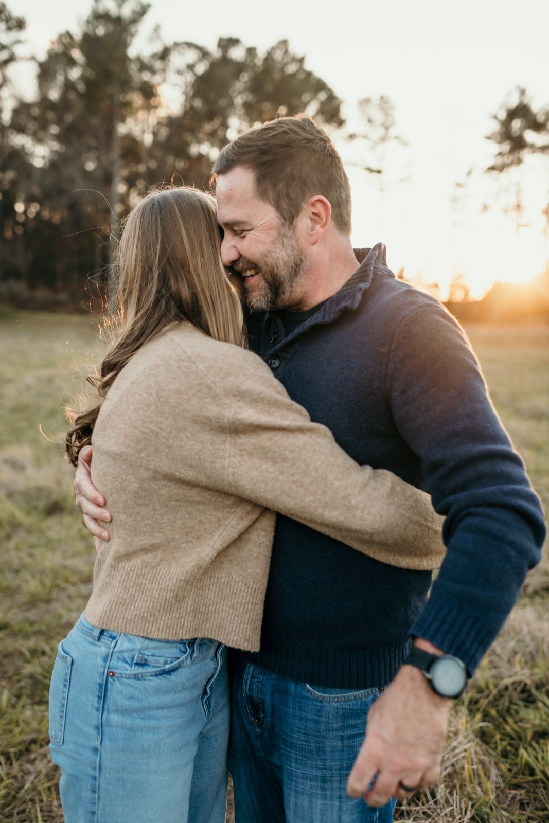 A couple embraces in a grassy field during golden hour sunset, wearing casual sweaters and jeans.