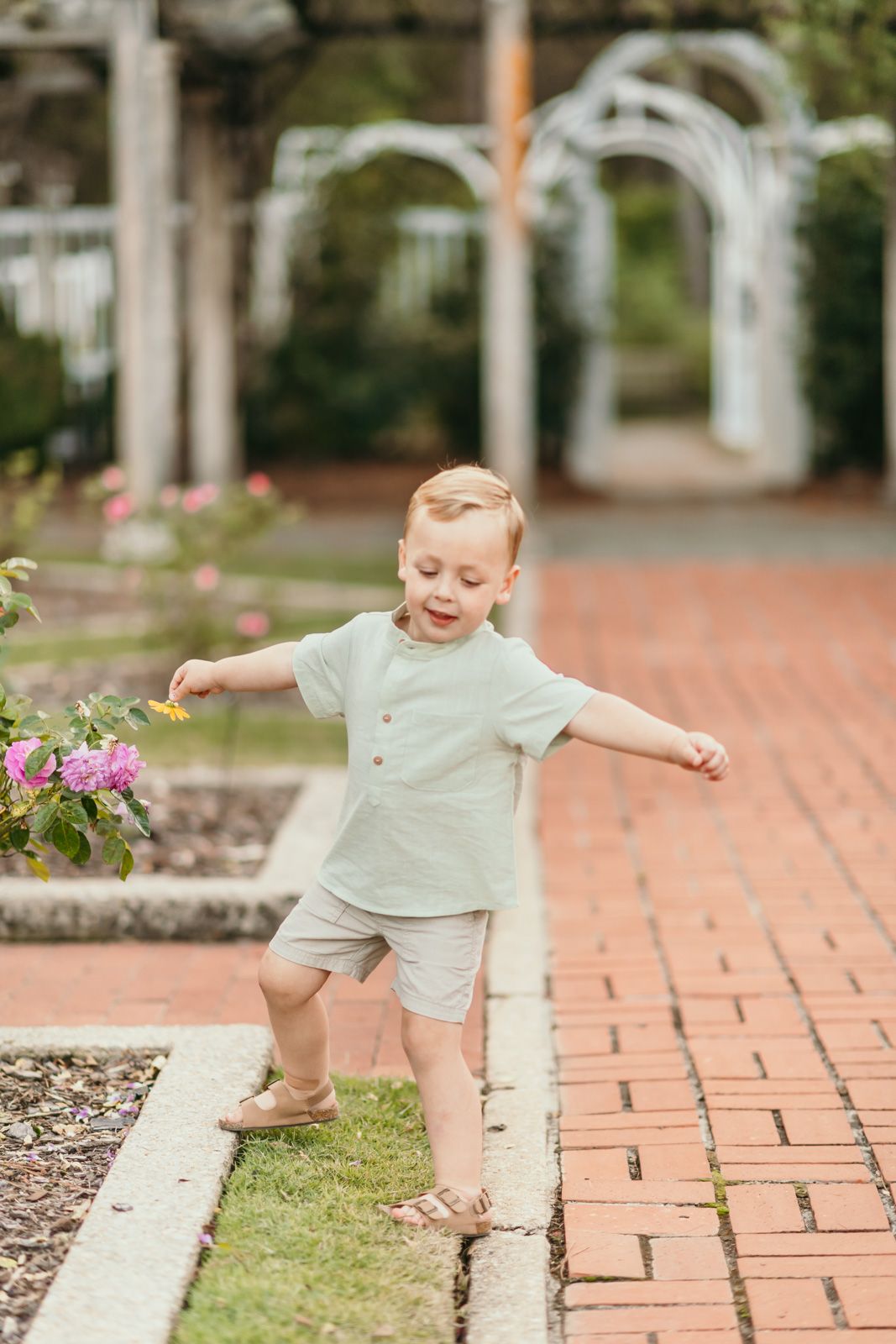 A toddler in a light colored outfit plays along a brick pathway in a garden with white arches in the background.