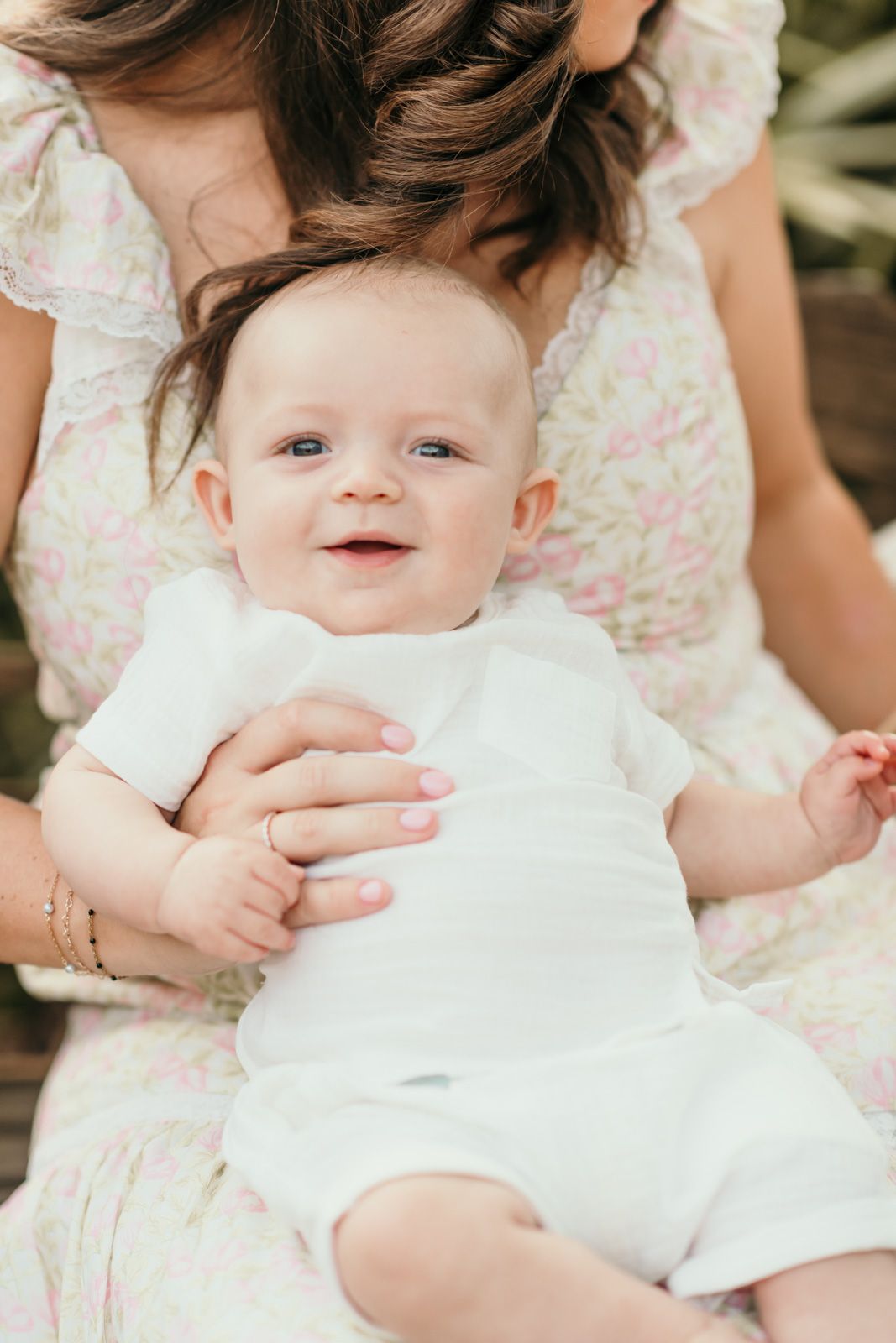 Baby in white outfit smiles while being held in floral setting.