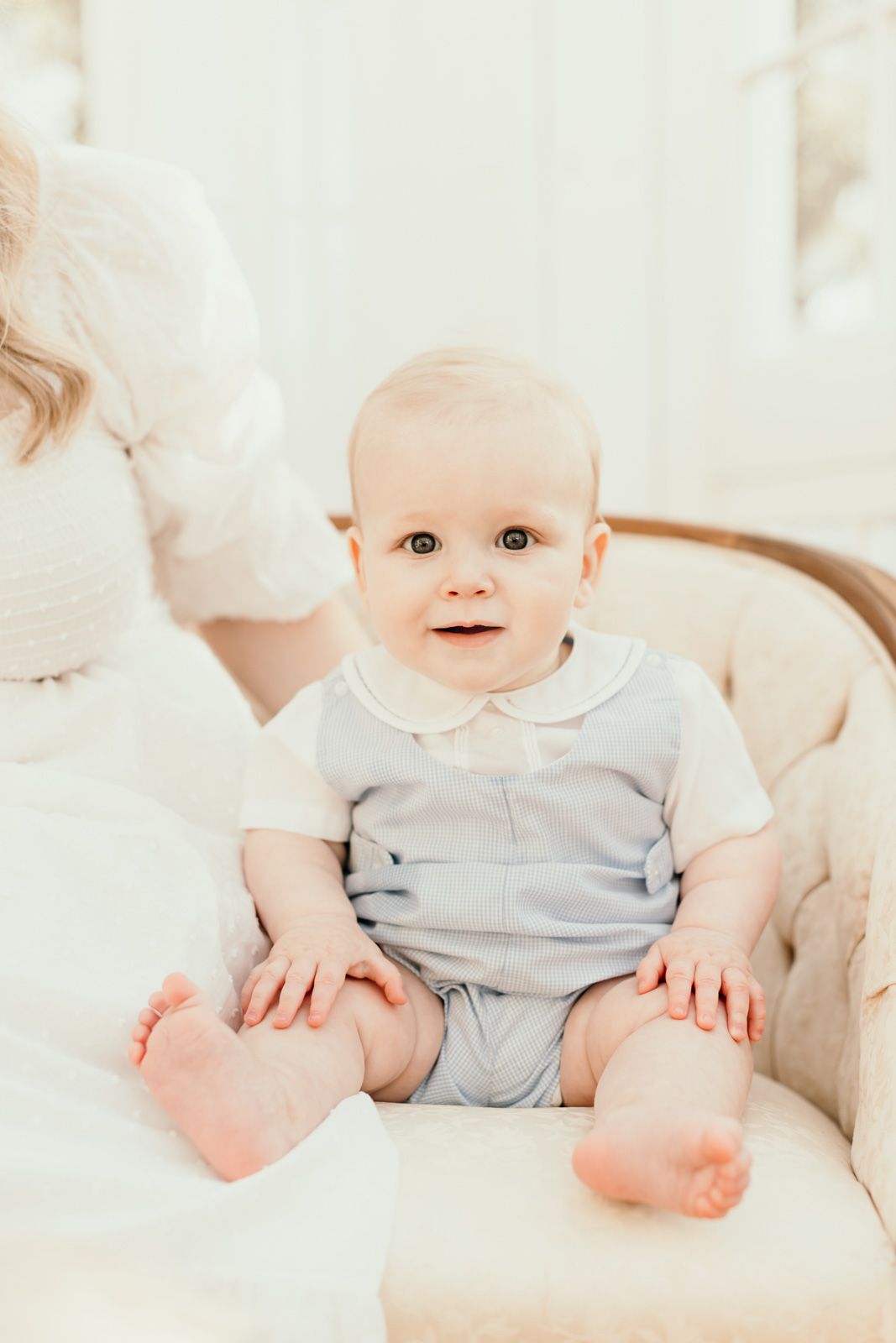 Sweet baby in light gray romper sits on plush white couch with bare feet showing.