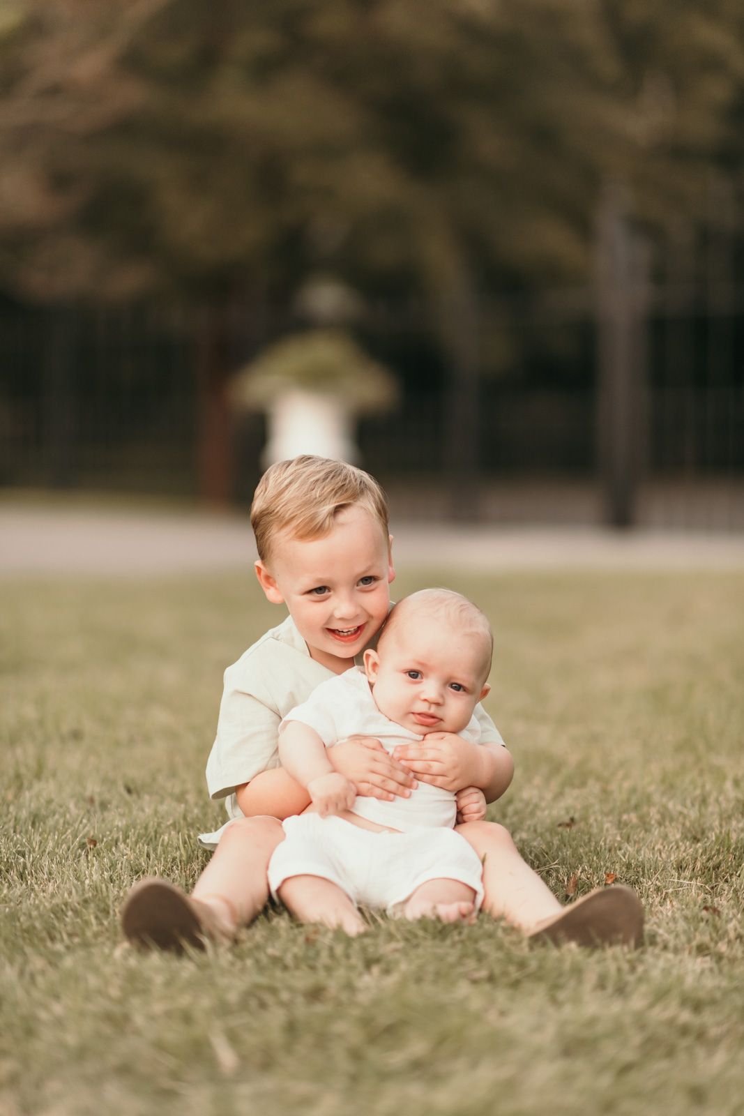 Two young children in white outfits sit together on grass outdoors while sharing a sweet moment.