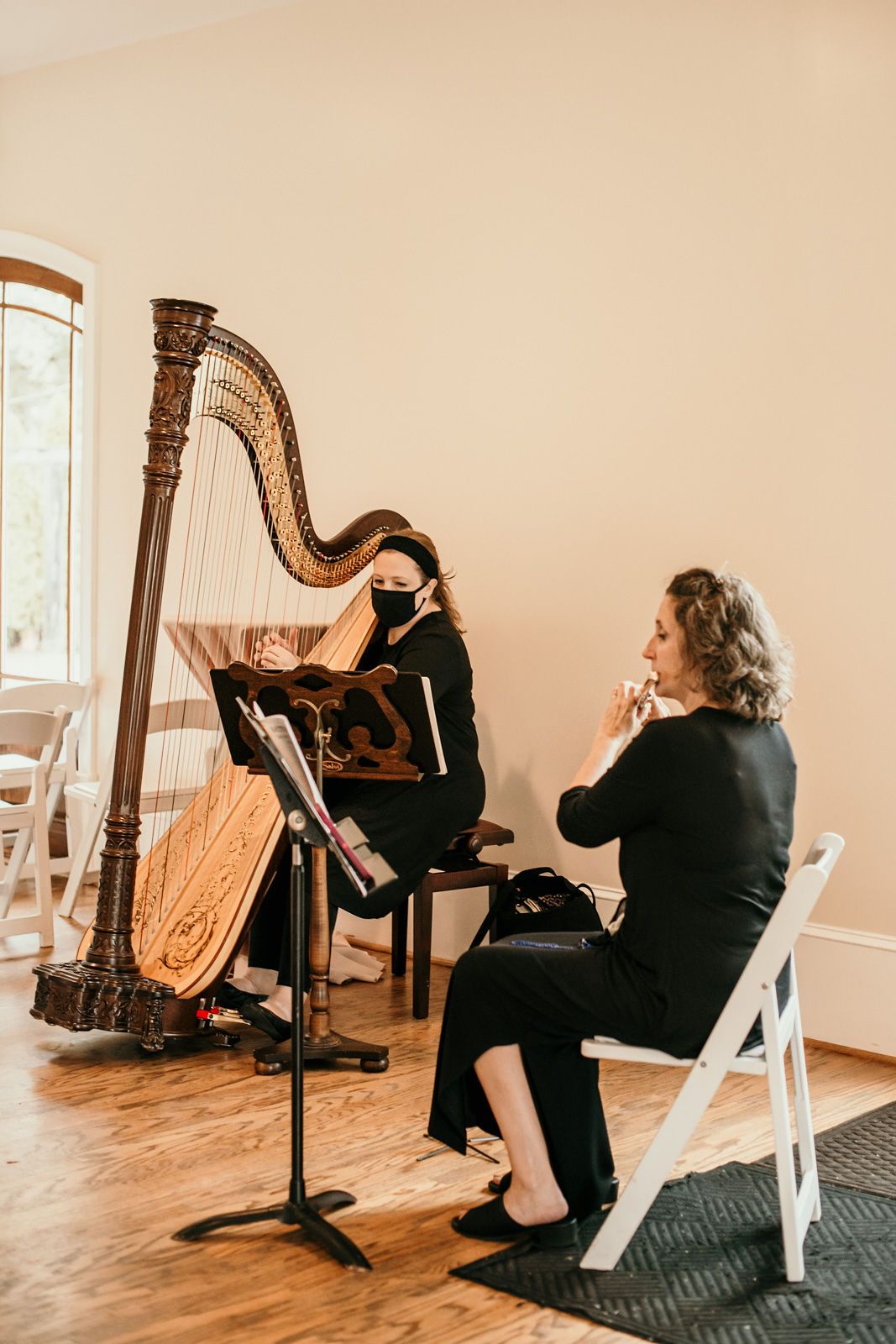 A harpist in black attire performs on an ornate wooden harp in a bright room with white walls.