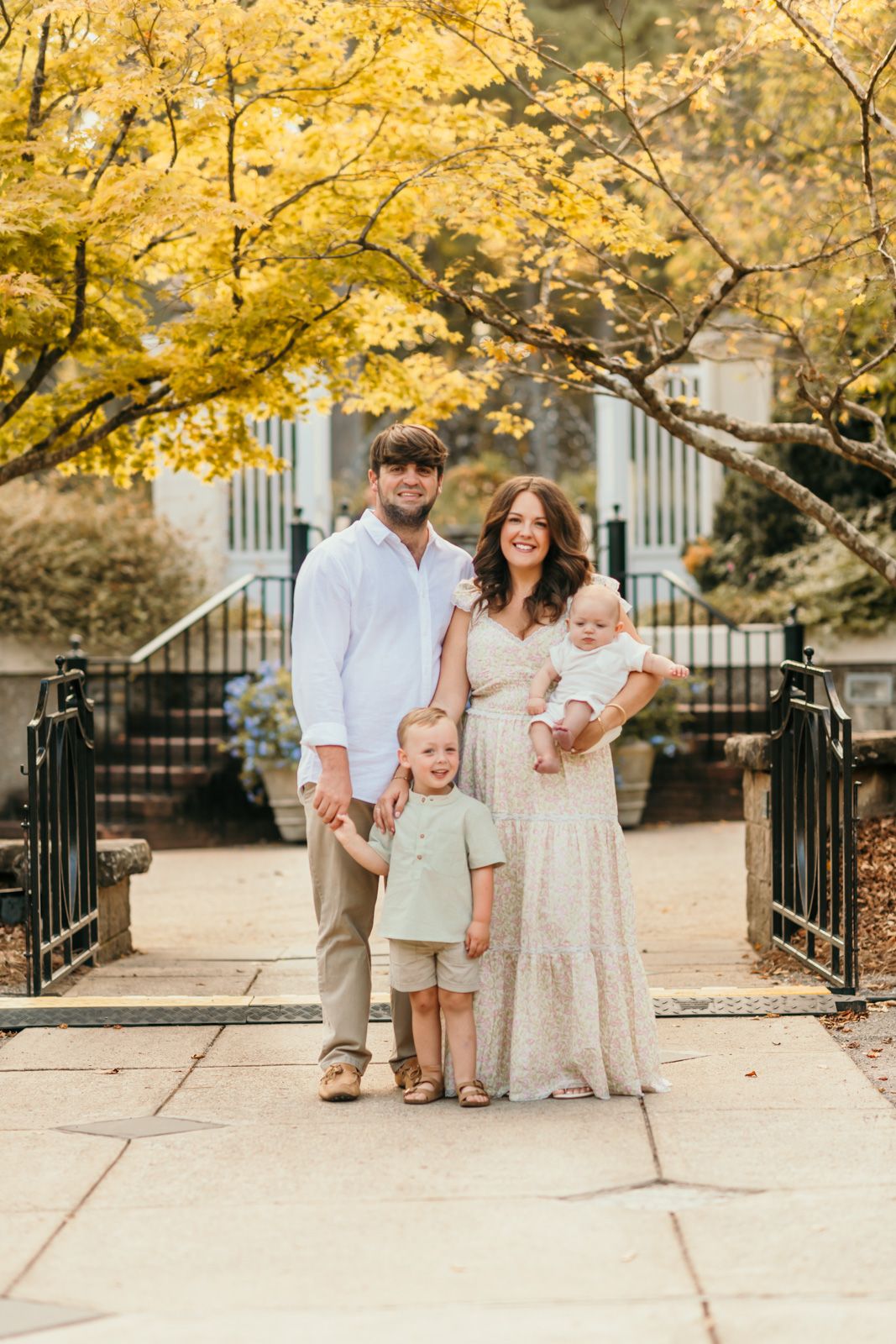 Family poses together on stone walkway under golden autumn trees in coordinated neutral outfits.
