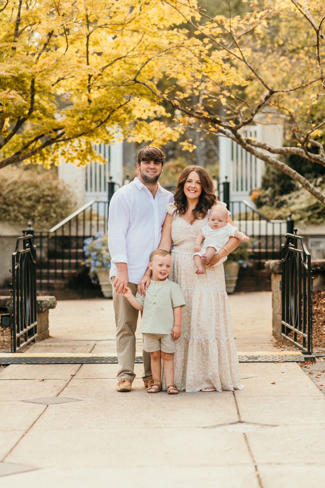 Family poses together under golden fall foliage near wrought iron fence.