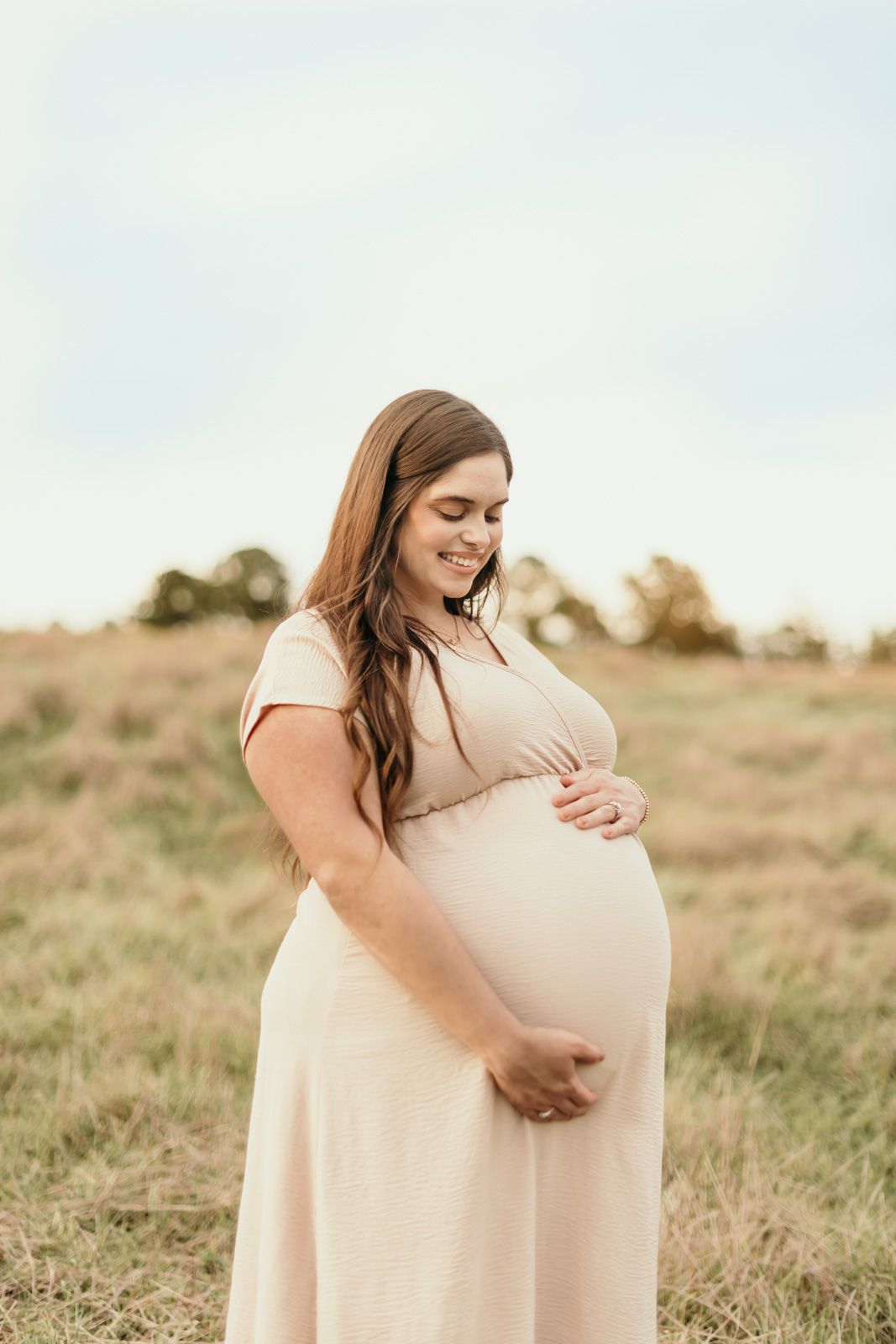 A pregnant woman in a cream dress stands in a grassy field during a maternity photoshoot at sunset.