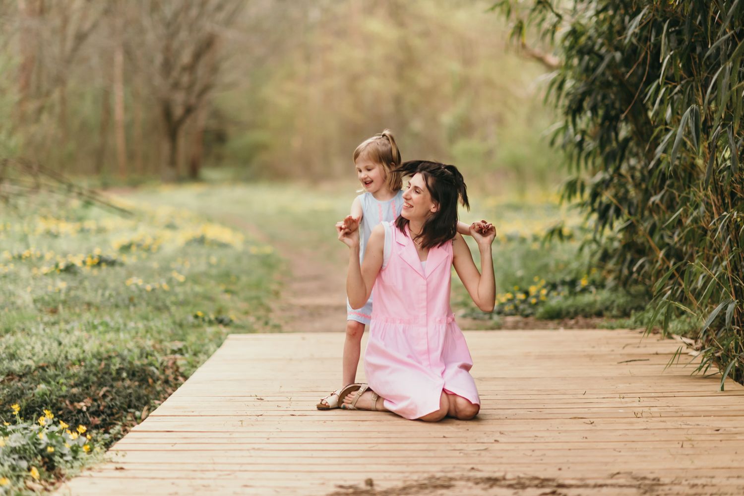 Two children in light summer clothing play together on a wooden boardwalk surrounded by yellow wildflowers and greenery.