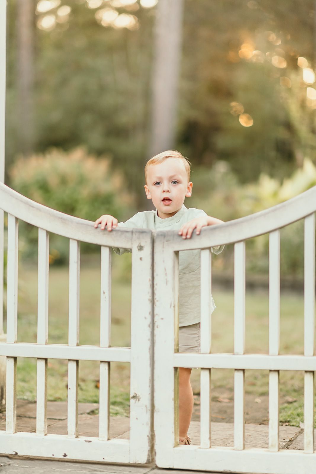 A toddler stands behind a white wooden fence in a garden setting with soft bokeh lighting in the background.