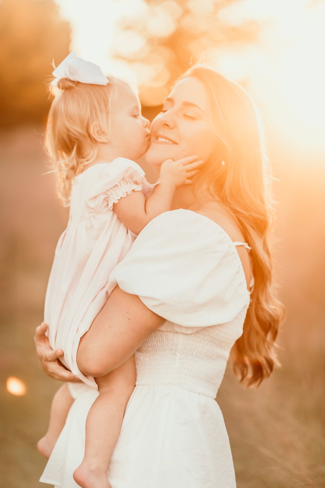 Mother and child embrace in soft golden sunlight wearing white dresses during outdoor portrait session.