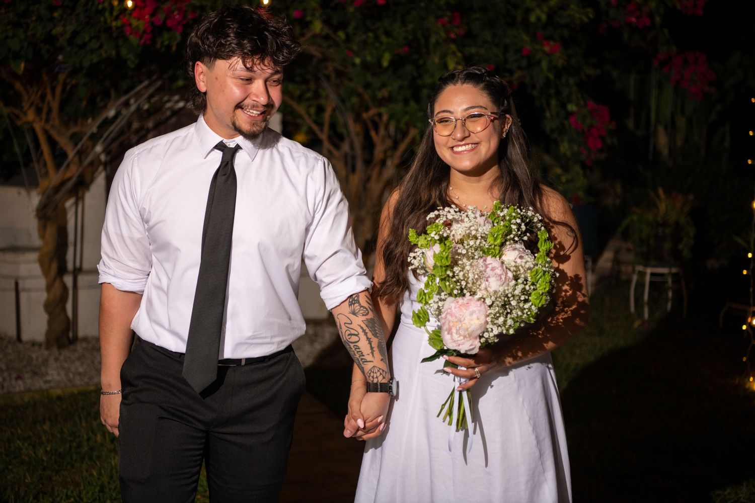 A couple in wedding attire holds hands while walking outdoors at night with a bouquet of white flowers. Wedding photography.