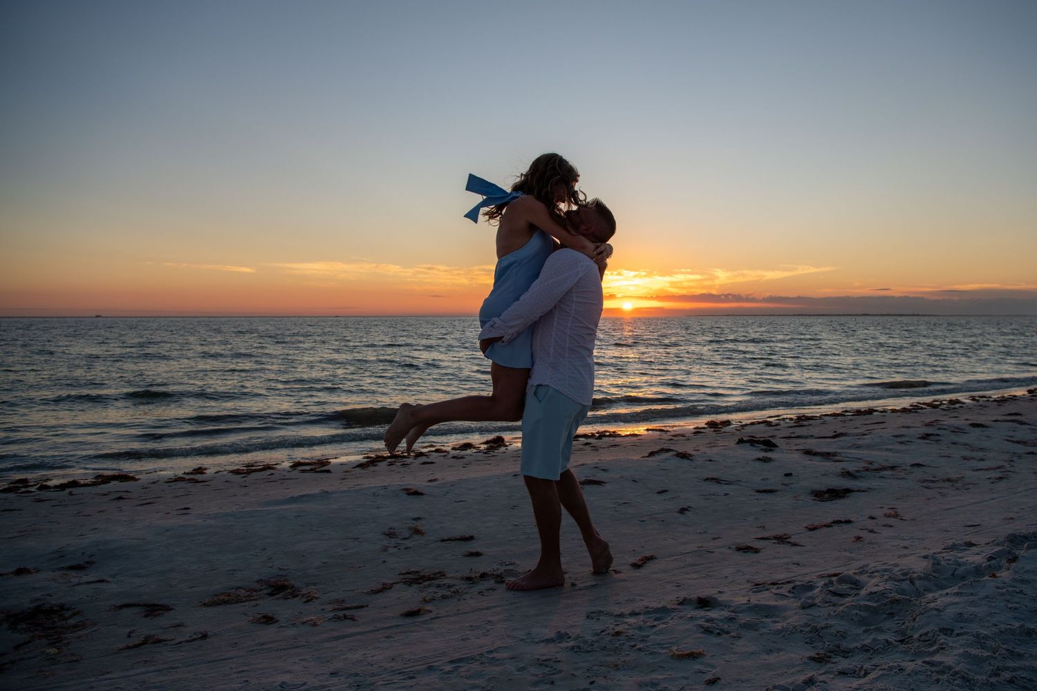 Silhouetted figures share tender moment on beach during sunset. Engagement photoshoot. Couples photos.