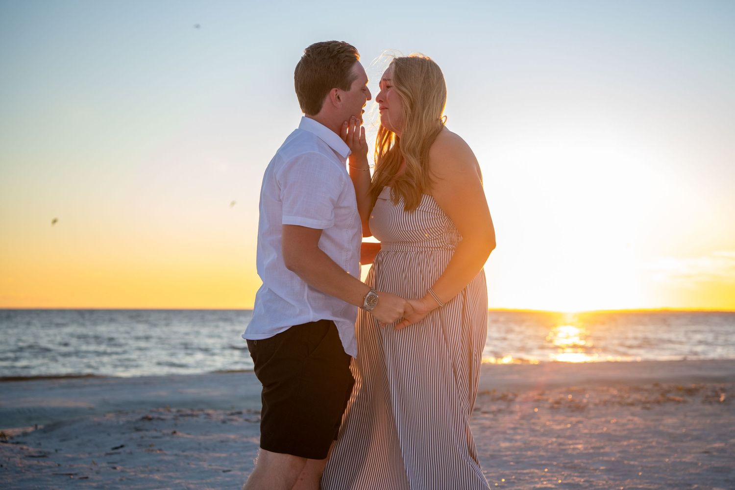 Couple embracing on sandy beach with dramatic sunset lighting and ocean backdrop.