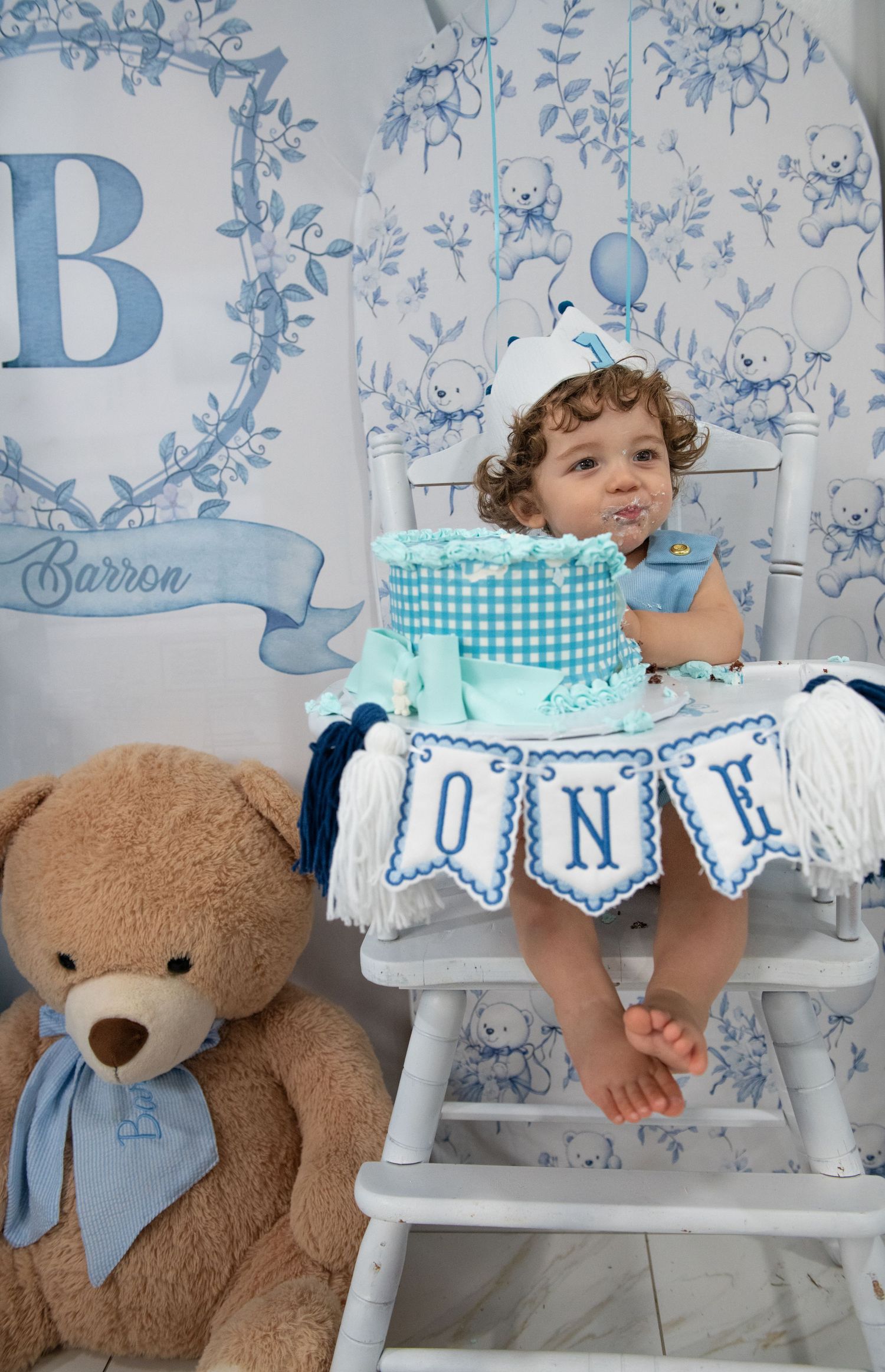First birthday celebration with teddy bear, white chair, and blue gingham cake.