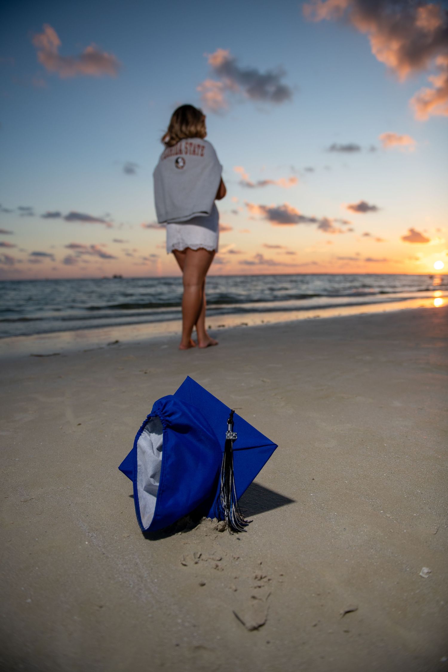 A blue graduation cap rests on a beach at sunset while a figure walks along the shoreline.
