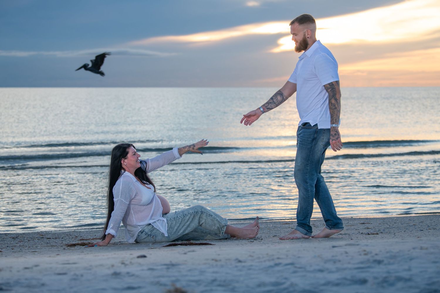 Couple enjoying romantic moments on beach with seabird flying overhead at sunset. Maternity photoshoot.