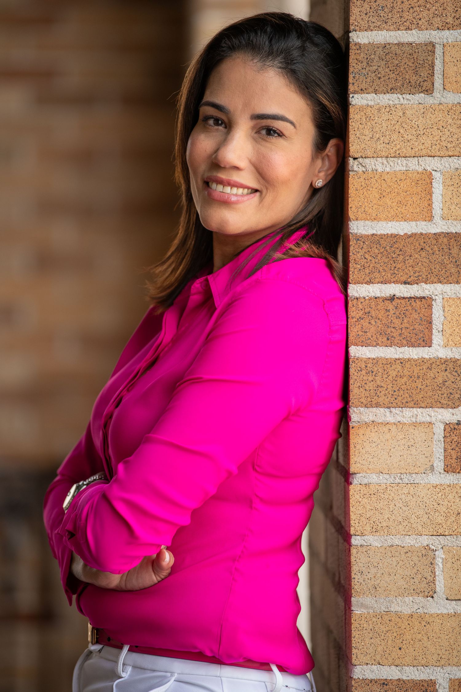 Business professional in bright pink blouse leaning against brick column with crossed arms.