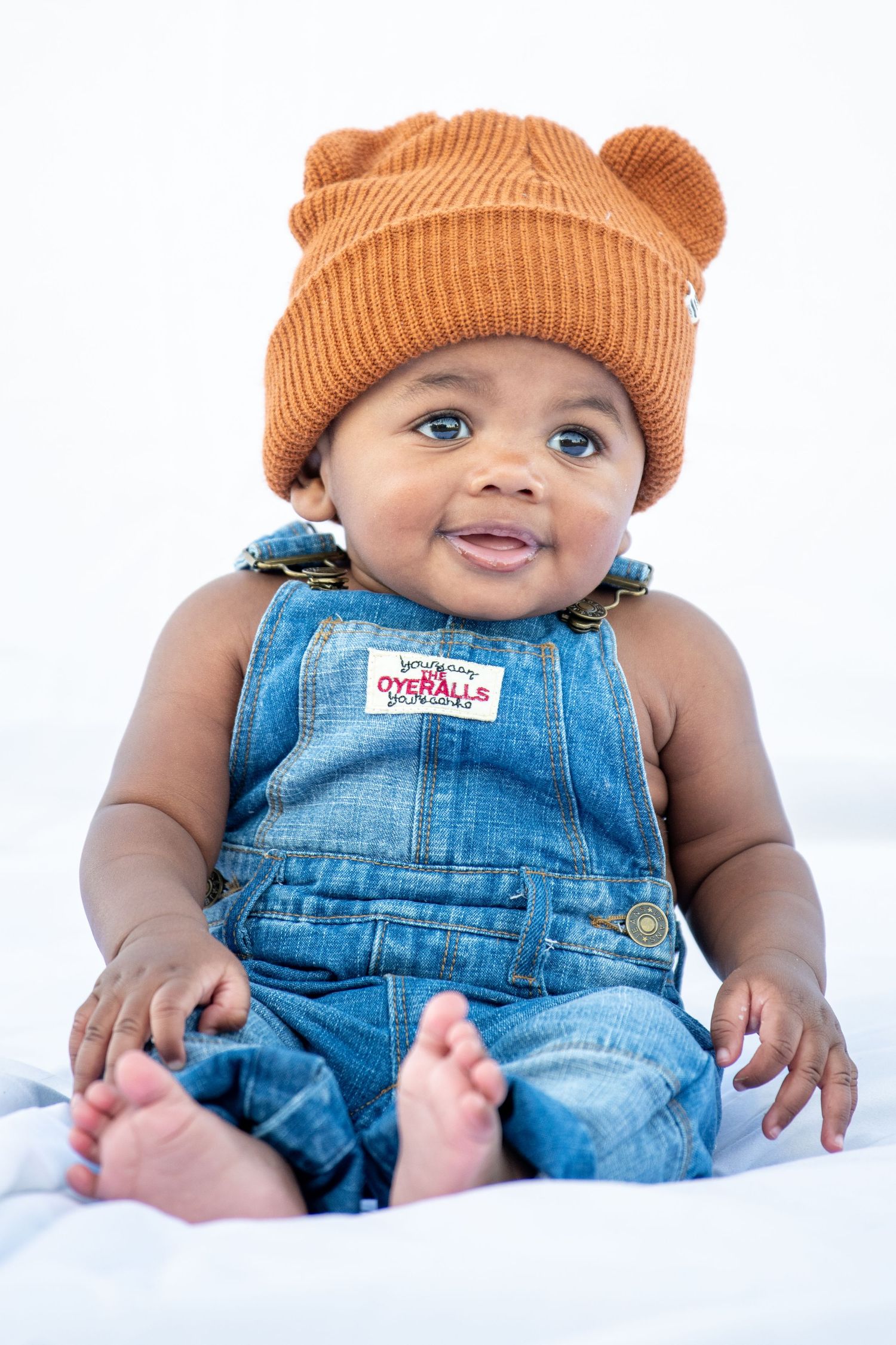 Infant in orange beanie and denim overalls poses on white backdrop.