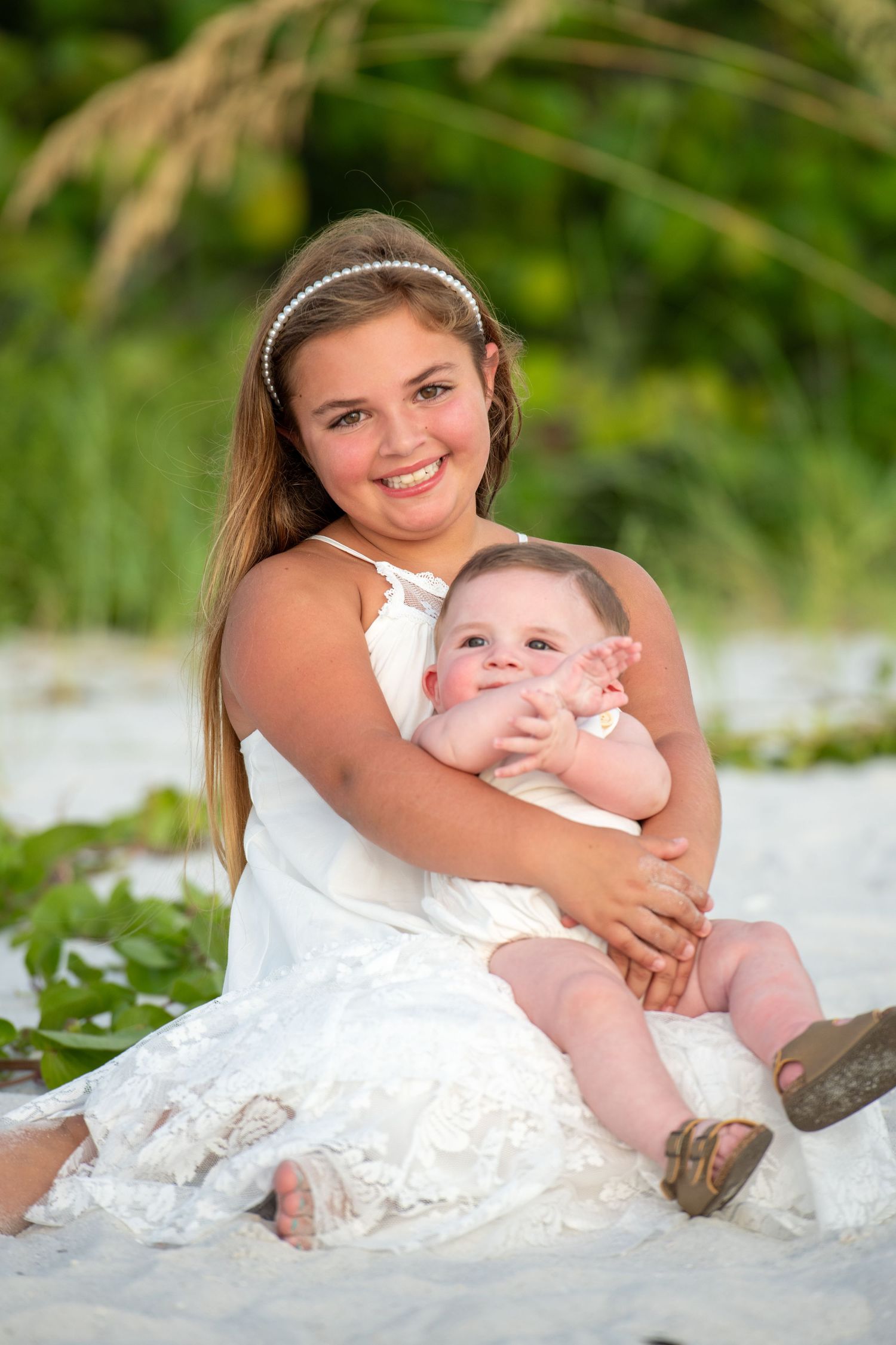 Young girl in white dress sitting on beach sand with baby during golden hour.