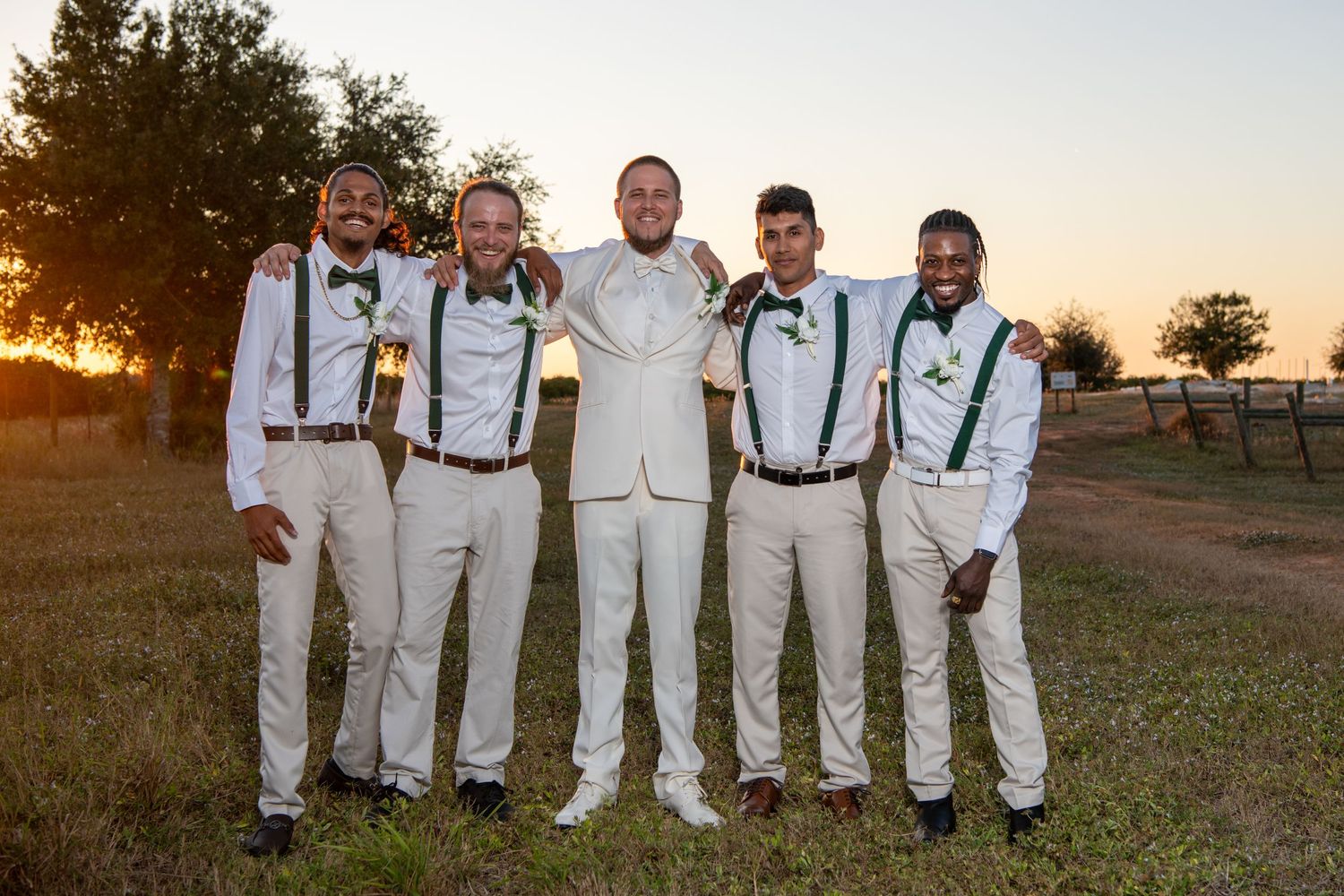 Groomsmen in white suits and suspenders pose together during outdoor sunset wedding. Wedding photography.