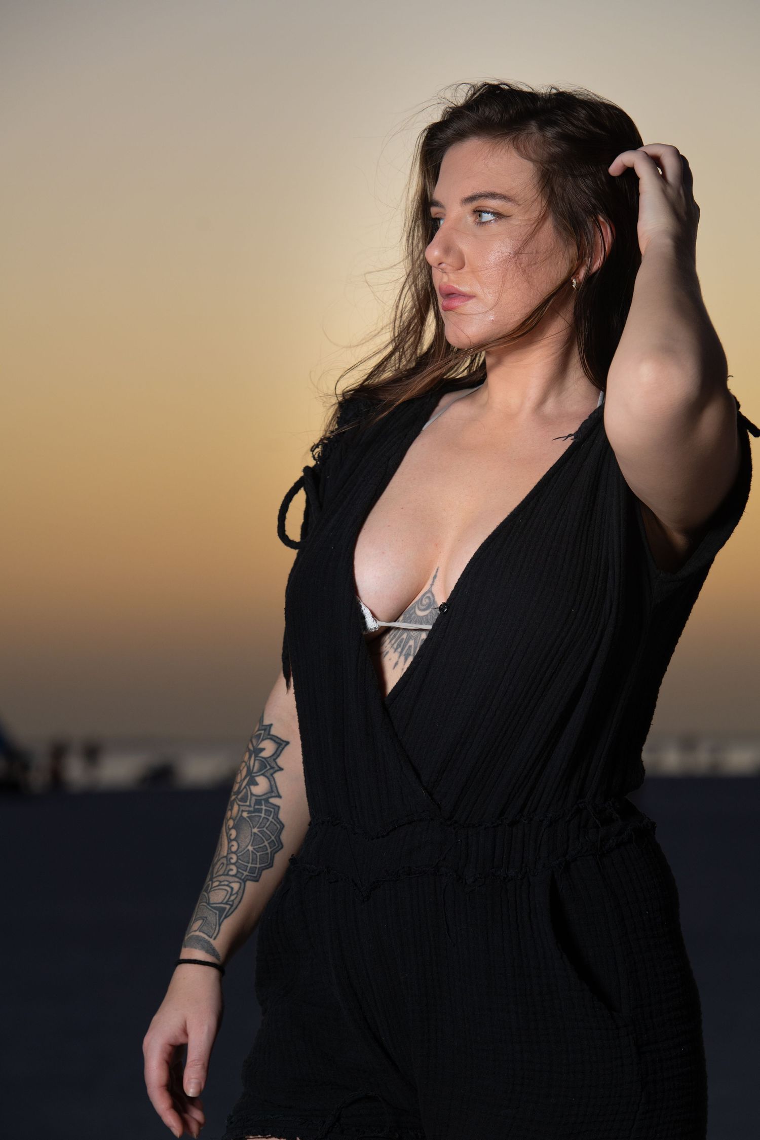 A woman in a black dress poses thoughtfully against a sunset sky backdrop.