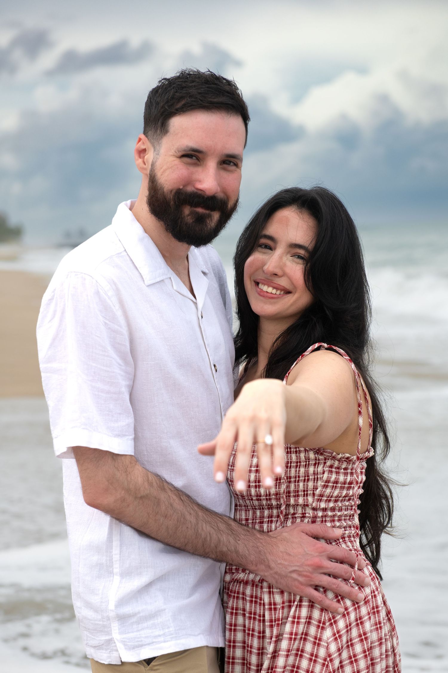 A couple embraces on a cloudy beach showing off an engagement ring.