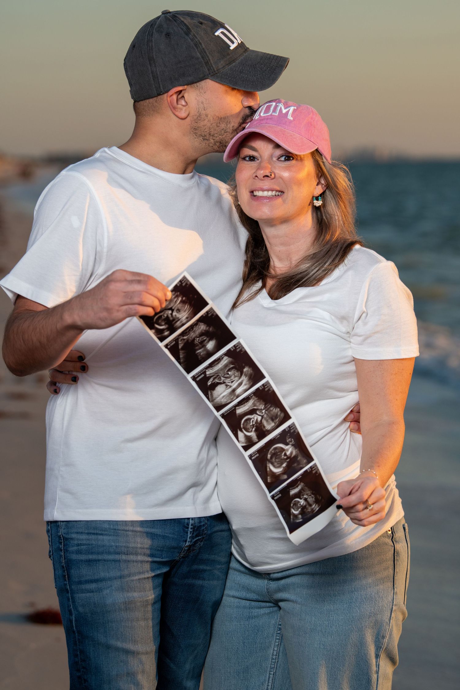 An expectant couple holding ultrasound photos while posing together on the beach at sunset. Maternity photoshoot. Couples photos.