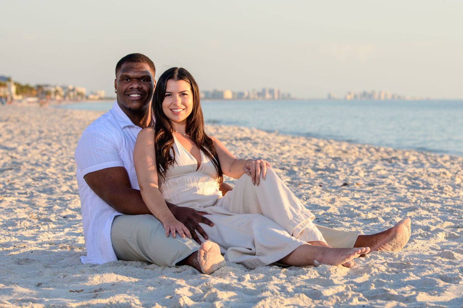 Couple relaxing on sandy beach with ocean and city skyline in background. Engagement photoshoot. Couples photos.