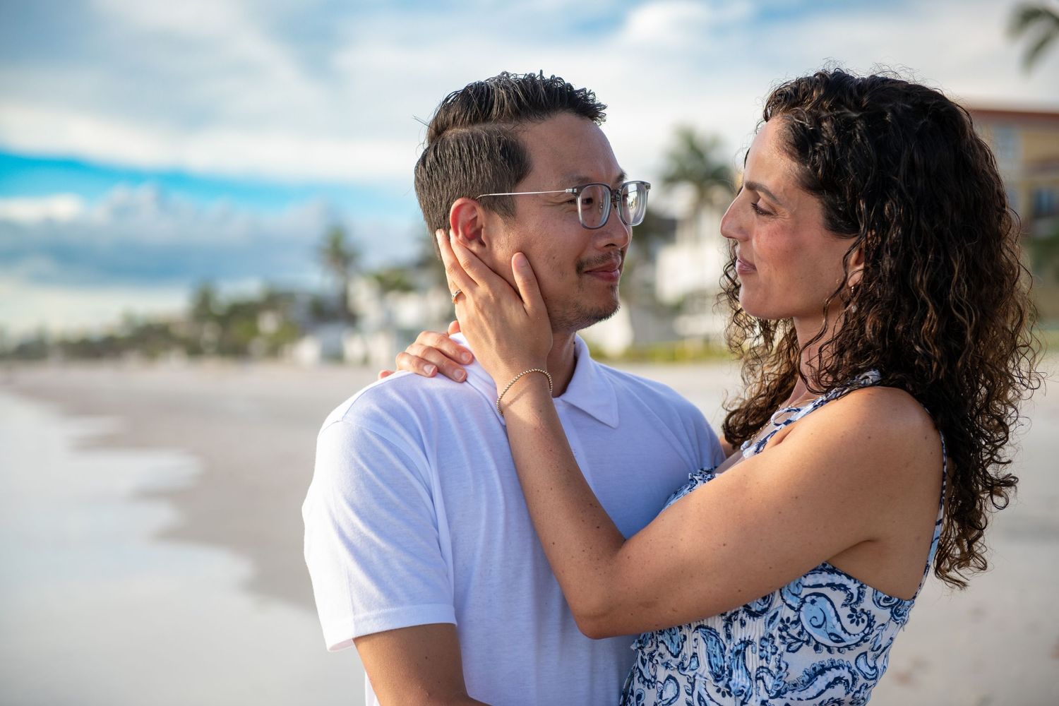 A romantic couple embraces on a tropical beach with palm trees and buildings in the background.
