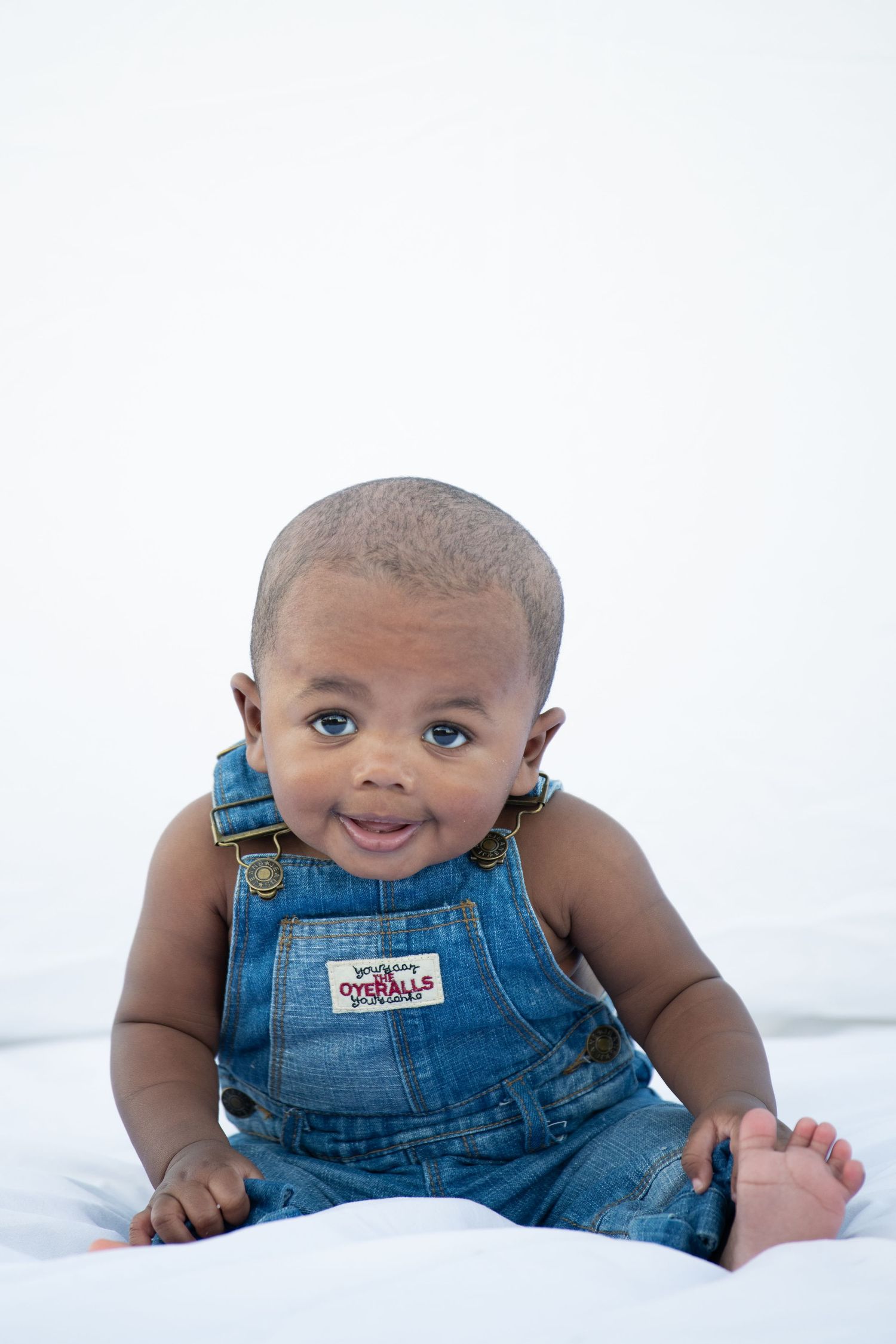 Baby in blue denim overalls smiles while lying on white surface.