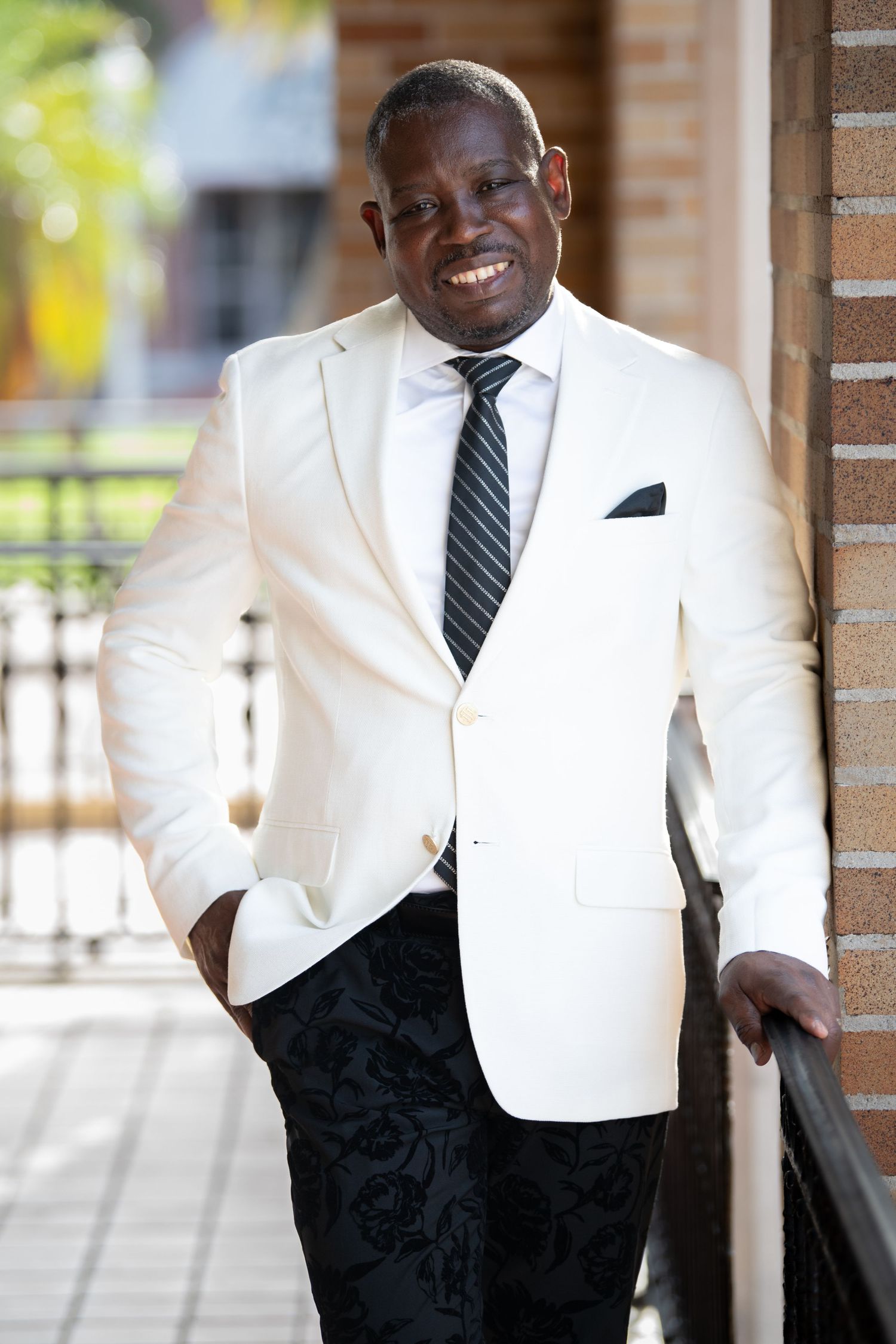 Stylish individual in white blazer and dark pants posing confidently by brick columns.