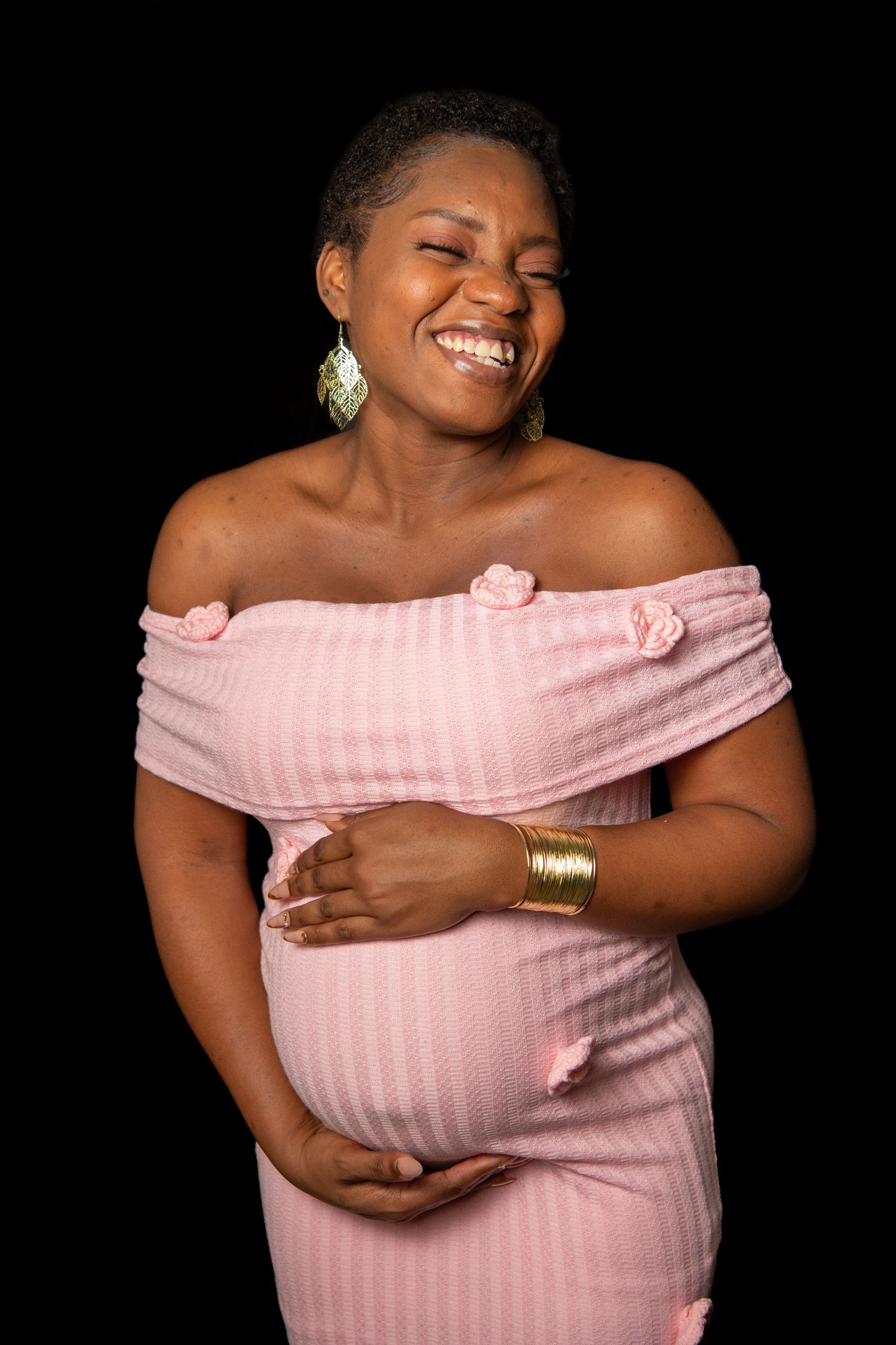 Maternity portraits in a soft pink off-shoulder dress against black background. Maternity photoshoot.