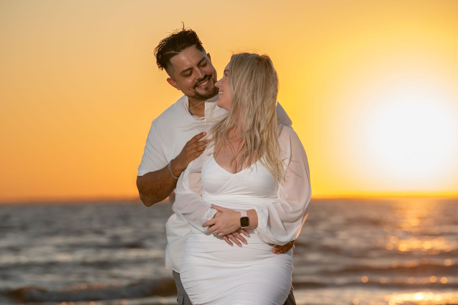 Romantic beach photoshoot during golden hour with couple in white outfits by the water.