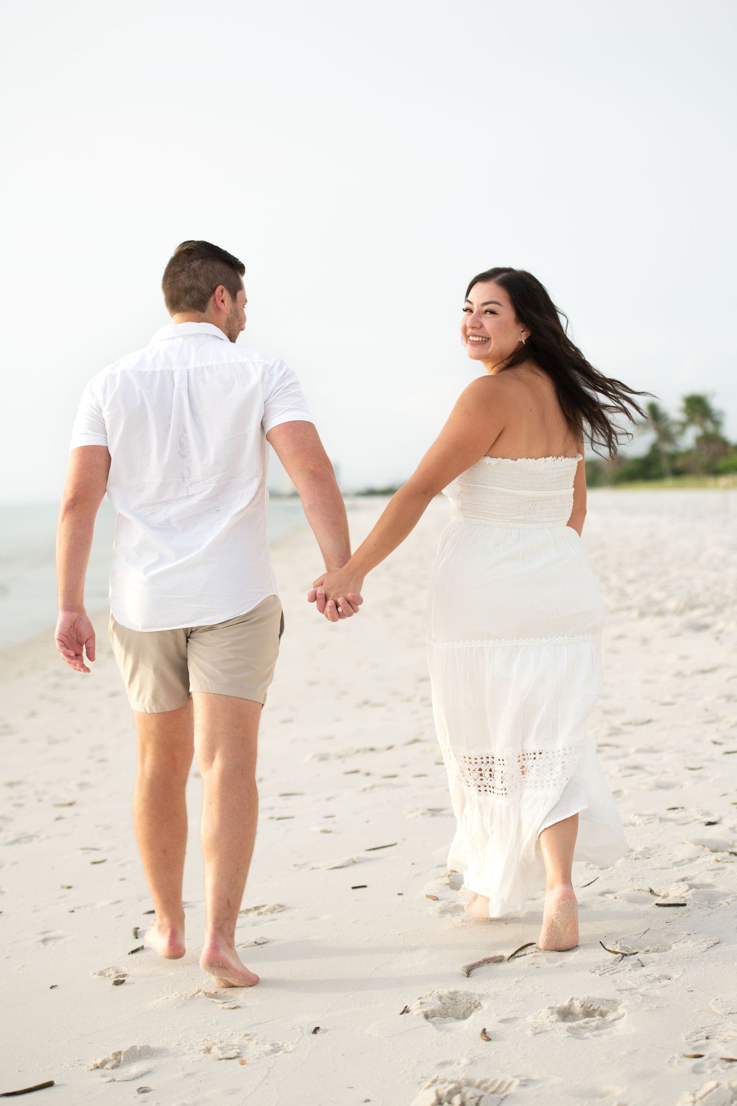 Couple walking hand in hand on white sandy beach wearing matching white outfits.